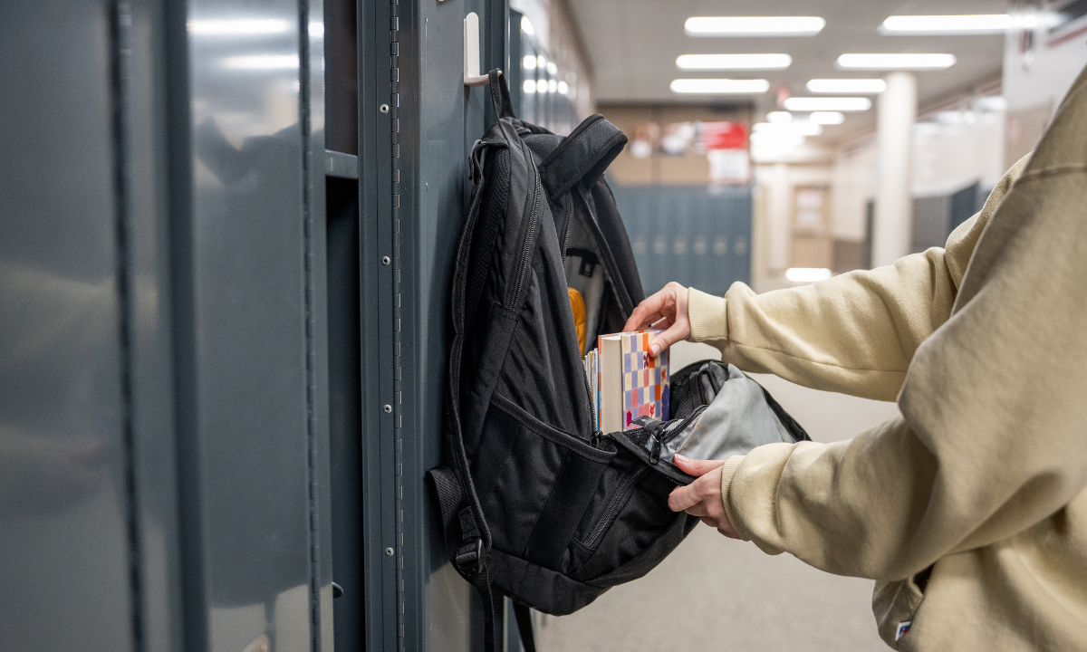 Black backpack hanging from a Gator magnetic hook on a steel locker as books are placed inside.
