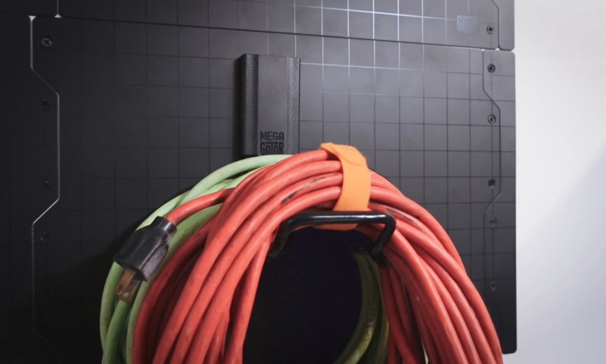 A black Gator Magnetics hook is mounted on a steel wall panel, holding tightly coiled red and green extension cords with an orange strap.
