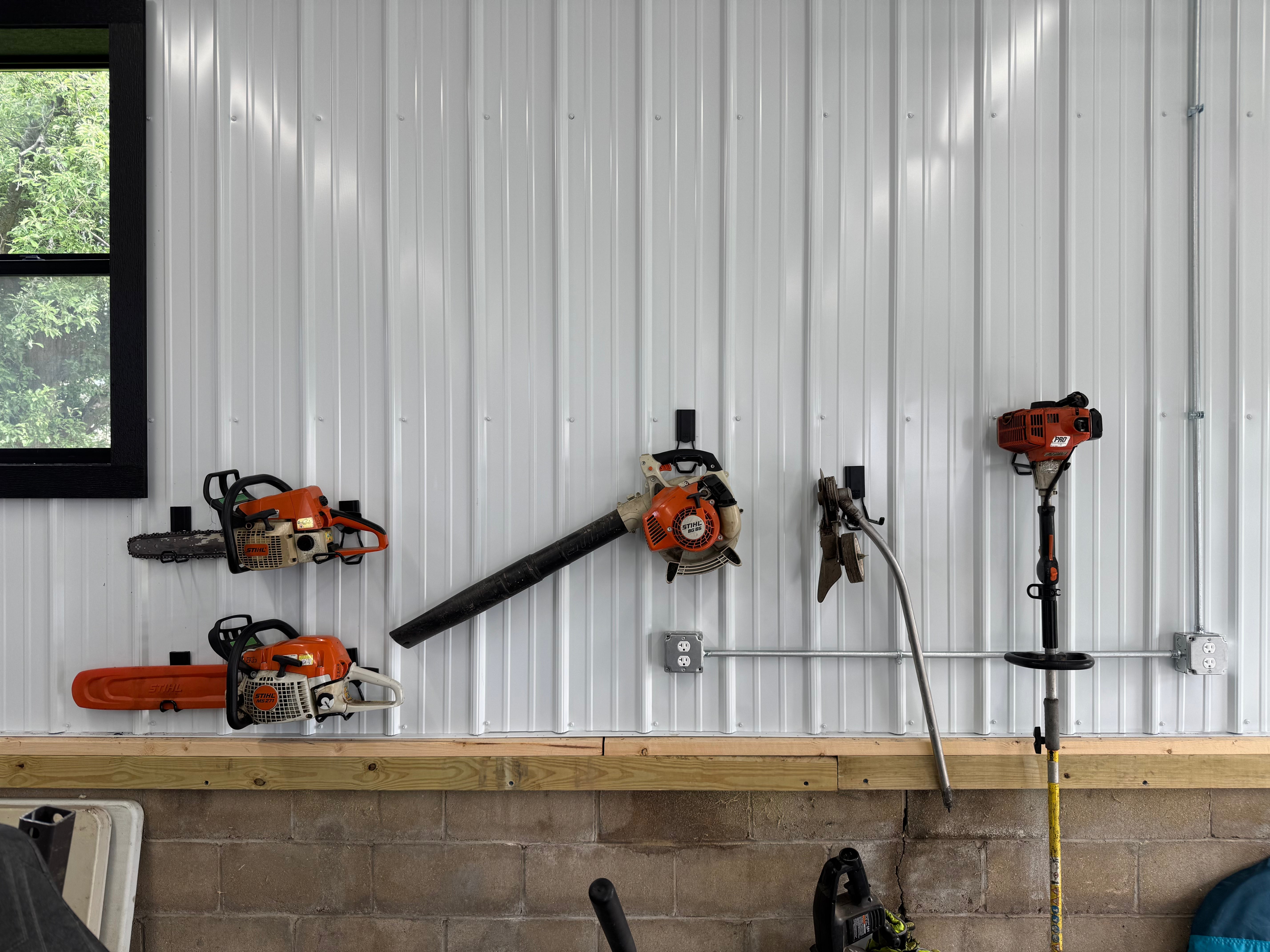 Gator magnetic hooks organize chainsaws, leaf blower, gloves and string trimmer on a white steel wall.