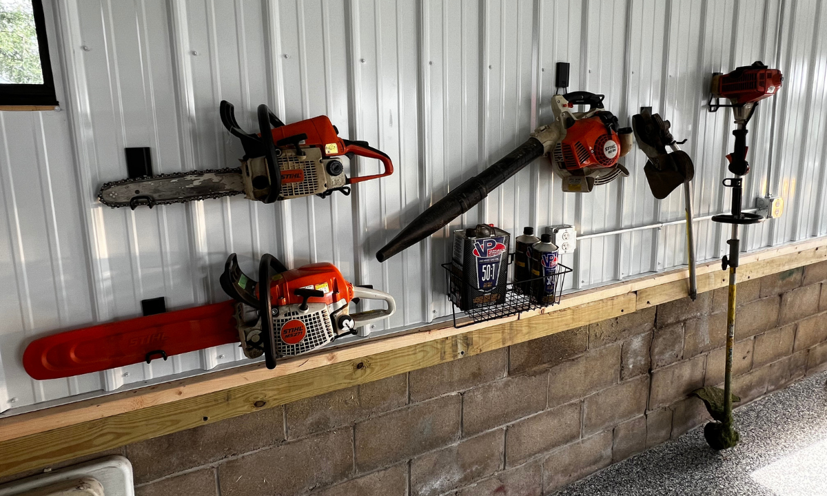 A chainsaw with blade guard and another chainsaw without guard hang on red magnetic hooks attached to a white steel pole barn wall panel.