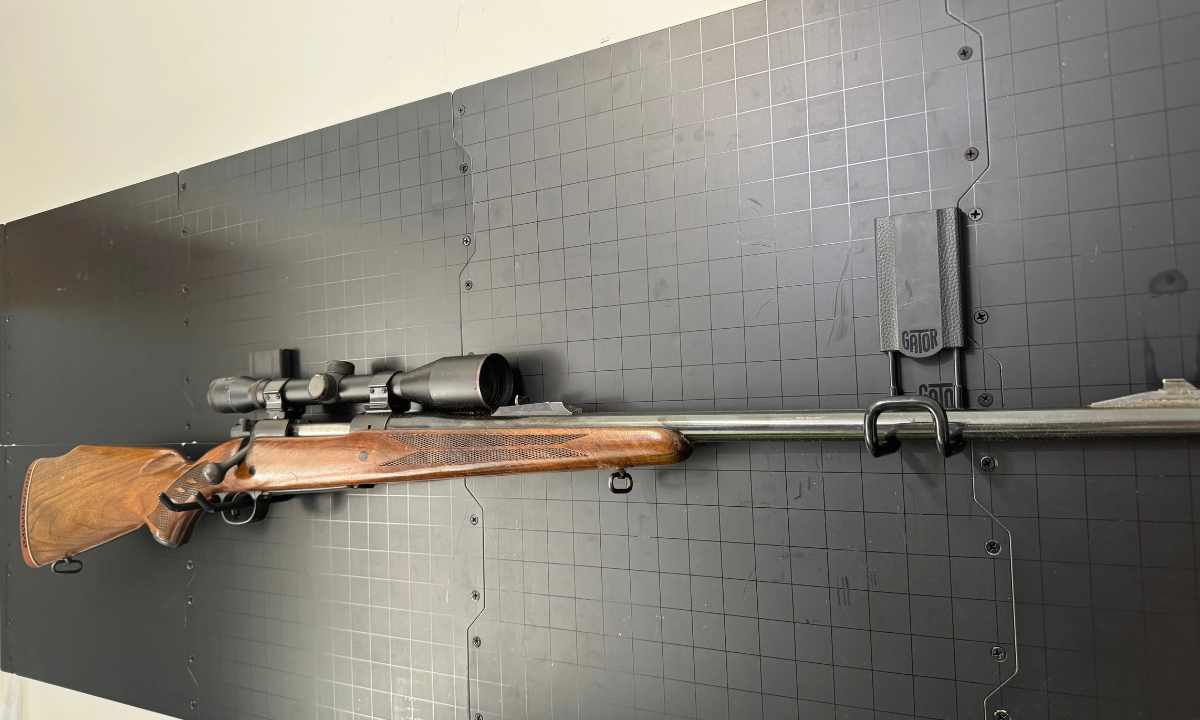 A hunting rifle with a scope is stored horizontally using a Gator Magnetics magnetic hook on a black steel wall panel system.