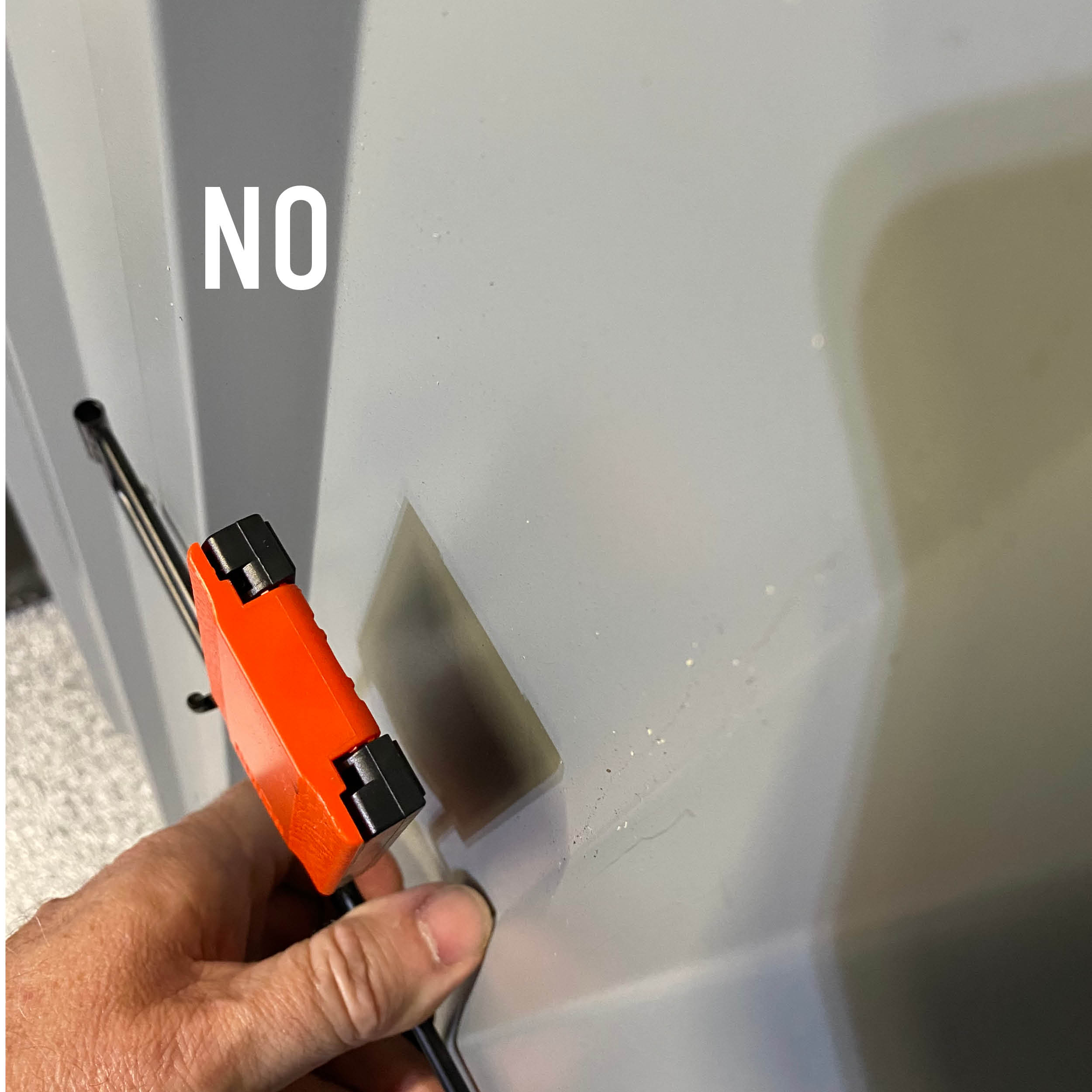 Hand holding a red magnetic hook with attached cables being positioned on a flat gray steel surface with residual adhesive residue and printed "NO" text, demonstrating a surface unsuitable for Gator Magnetics' towel baskets or hooks installation.