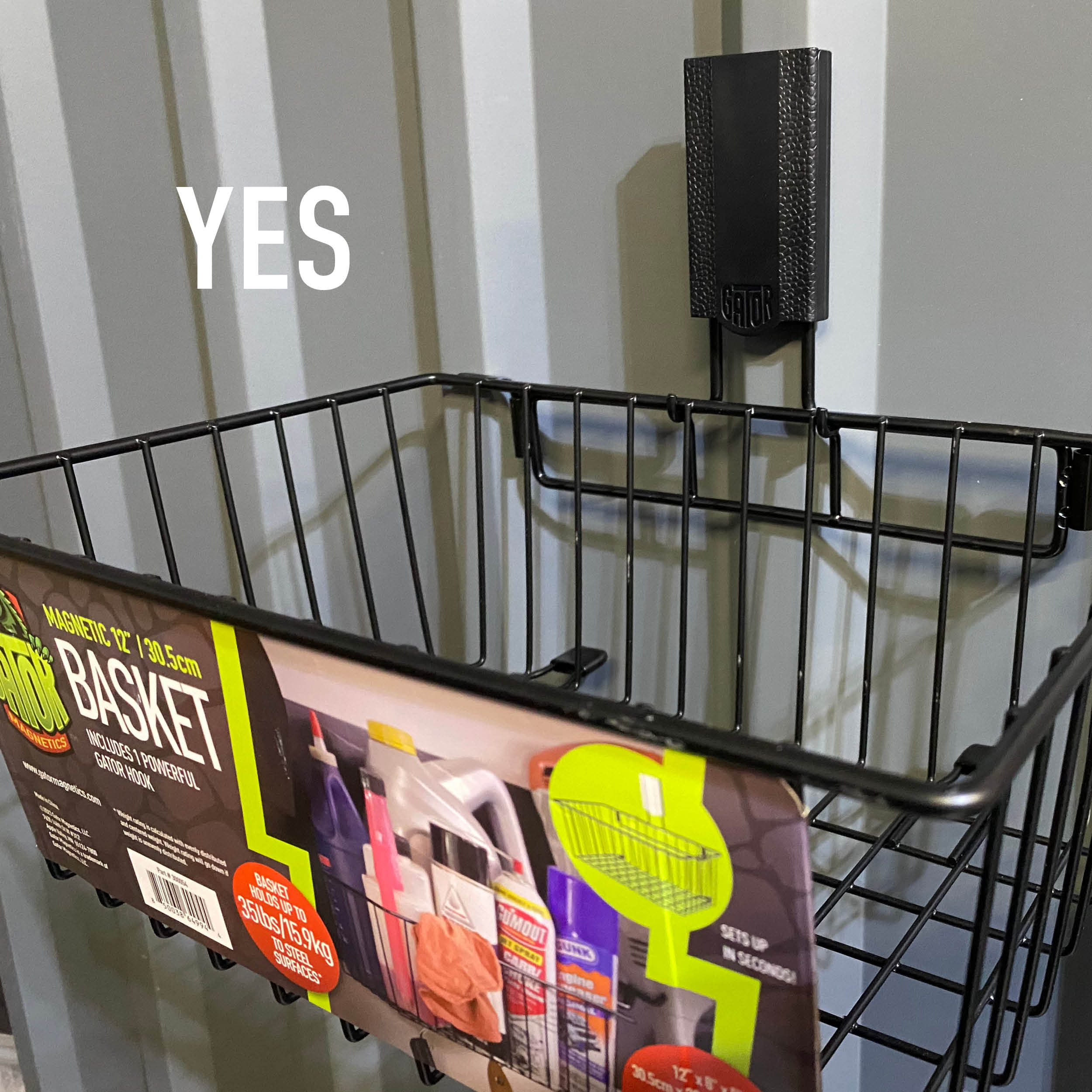 A black Gator Magnetics Magnetic Storage Basket is mounted on a flat steel pole barn steel wall panel, shown empty with packaging label visible, demonstrating a stylish and practical garage idea for organizing automotive fluids and cleaning supplies.