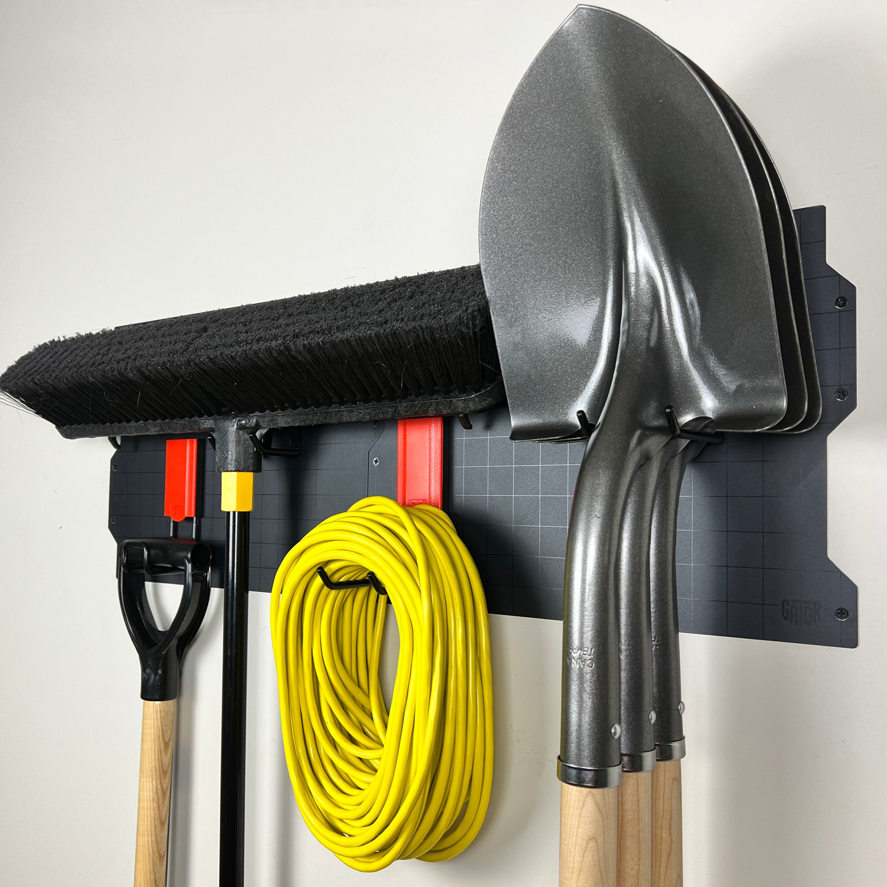 Three garden shovels, a broom, a coiled yellow extension cord, and a garden spade handle are hung on red and black gator magnet hooks attached to a black Gator Magnetics steel wall panel mounted on a flat steel surface.
