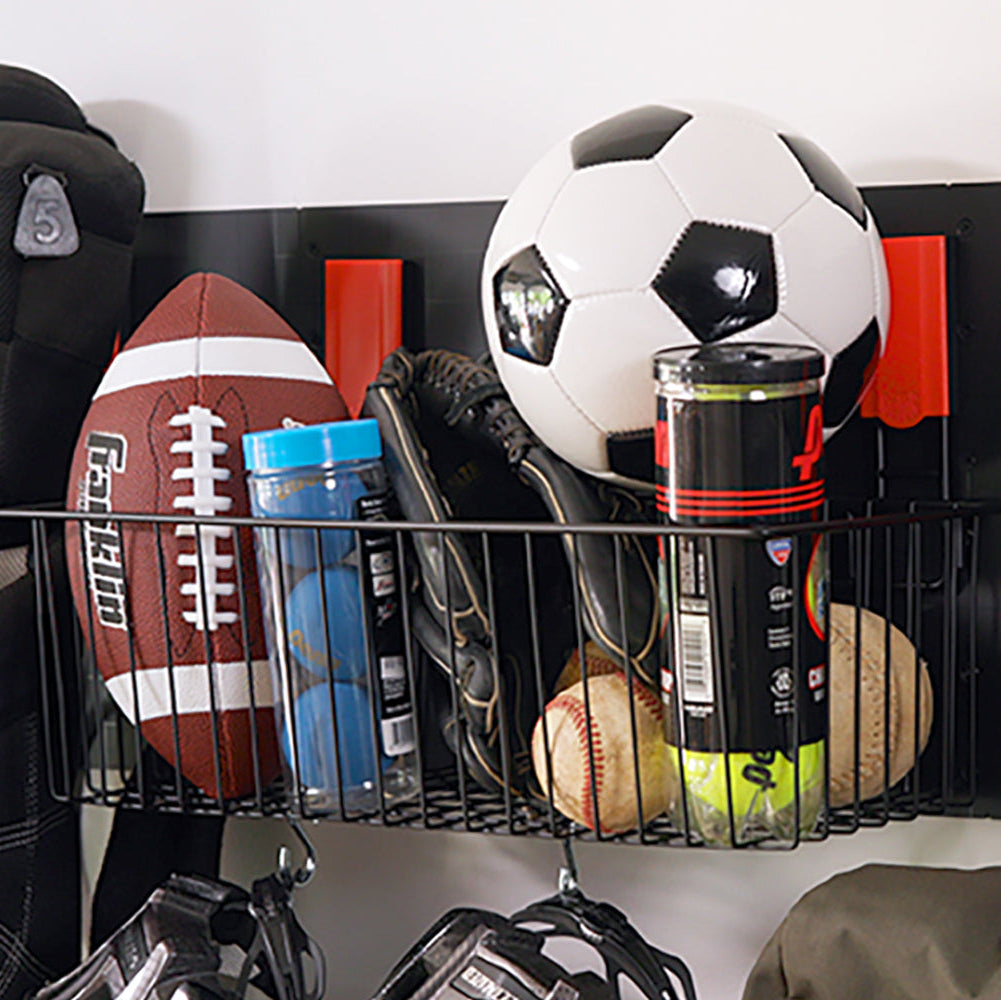 A black Gator Magnetics magnetic storage basket is mounted on a flat steel wall panel, holding a football, a soccer ball, baseball gloves, baseballs, tennis balls in a can, and a blue container, with red magnetic hooks attached to the panel underneath for additional hanging storage, showcasing practical sports gear organization including a magnet towel holder option.
