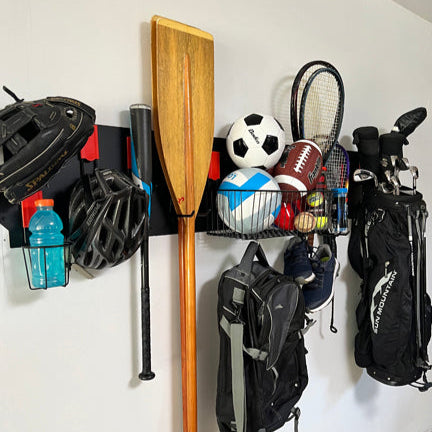 A black Gator Magnetics steel wall panel system mounted on a smooth steel wall stores multiple sports and outdoor items, including black and white soccer and footballs in magnetic storage baskets, a wooden canoe paddle, baseball bat, baseball glove and helmet hanging from red magnetic hooks, golf clubs and bags, tennis racket, black backpack, and a blue sports drink bottle, demonstrating versatile organization with magnetic oven mitts-compatible steel surfaces.