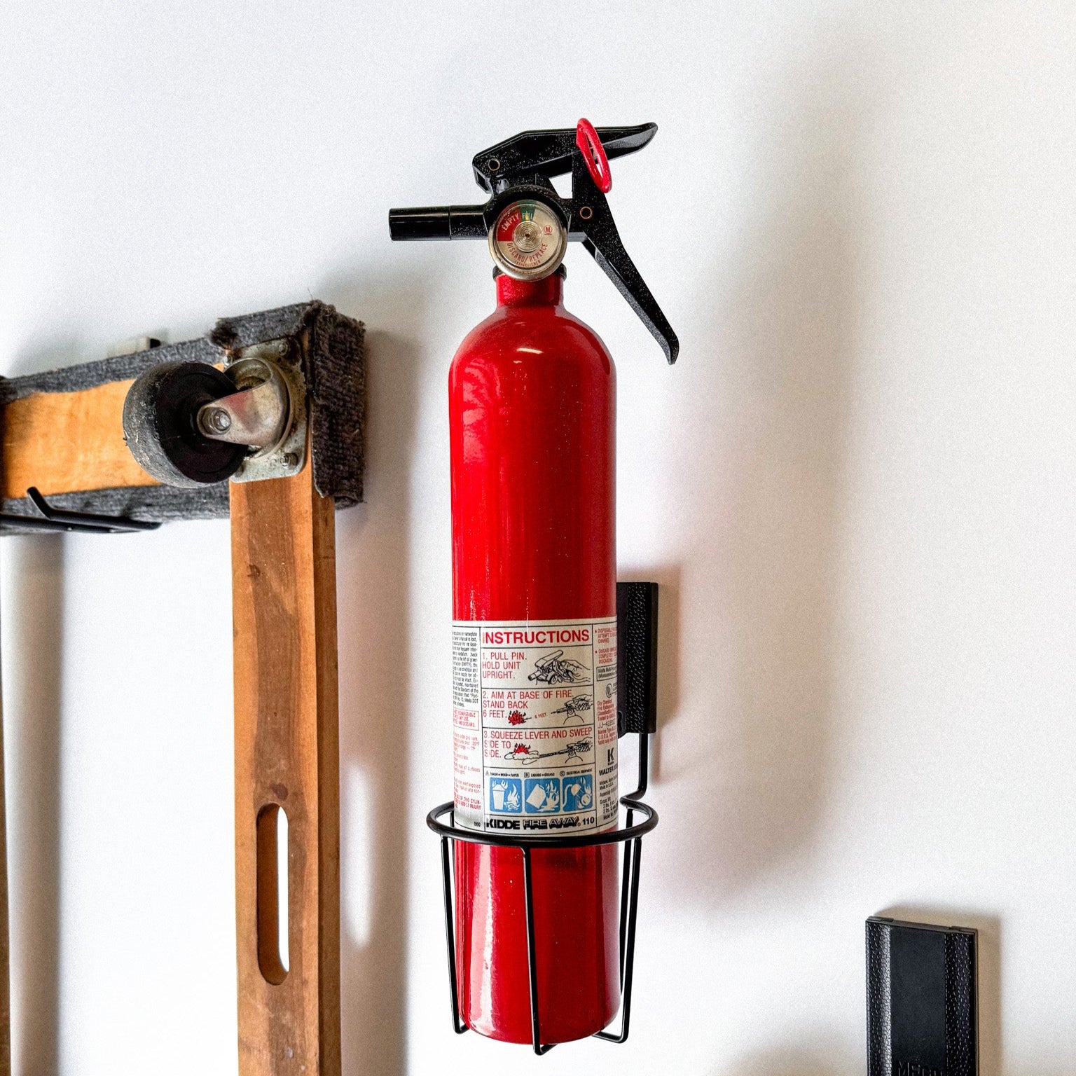 Gator Magnetics magnetic cup holder for garage storage and organization, mounted on a wall holding a red fire extinguisher, with a wooden furniture dolly and caster wheel in the background.