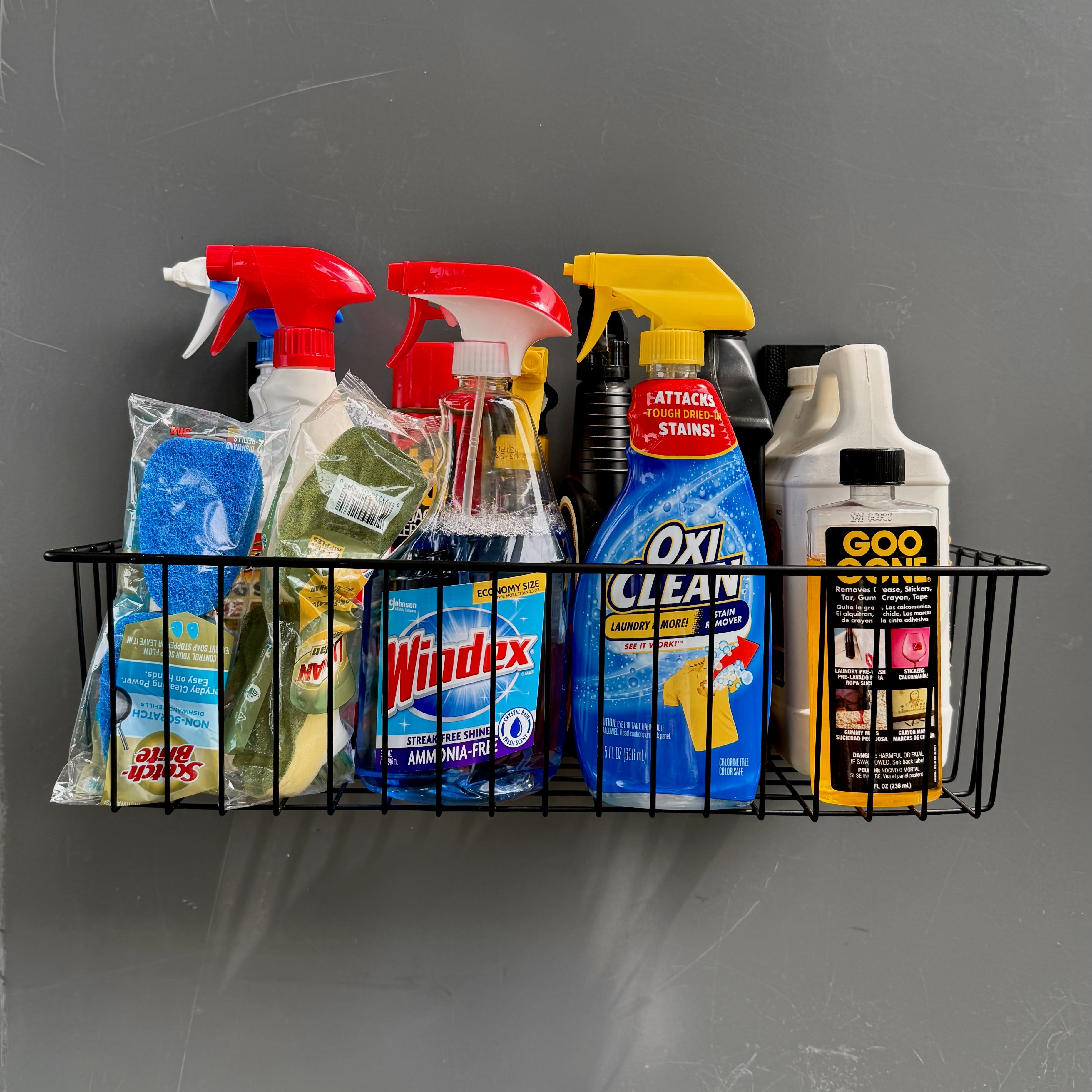 A Gator Magnetics magnetic wire basket organizer holds various cleaning supplies including spray bottles of Windex and OxiClean, sponges, and a Goo Gone bottle, all attached to a flat grey steel wall panel.