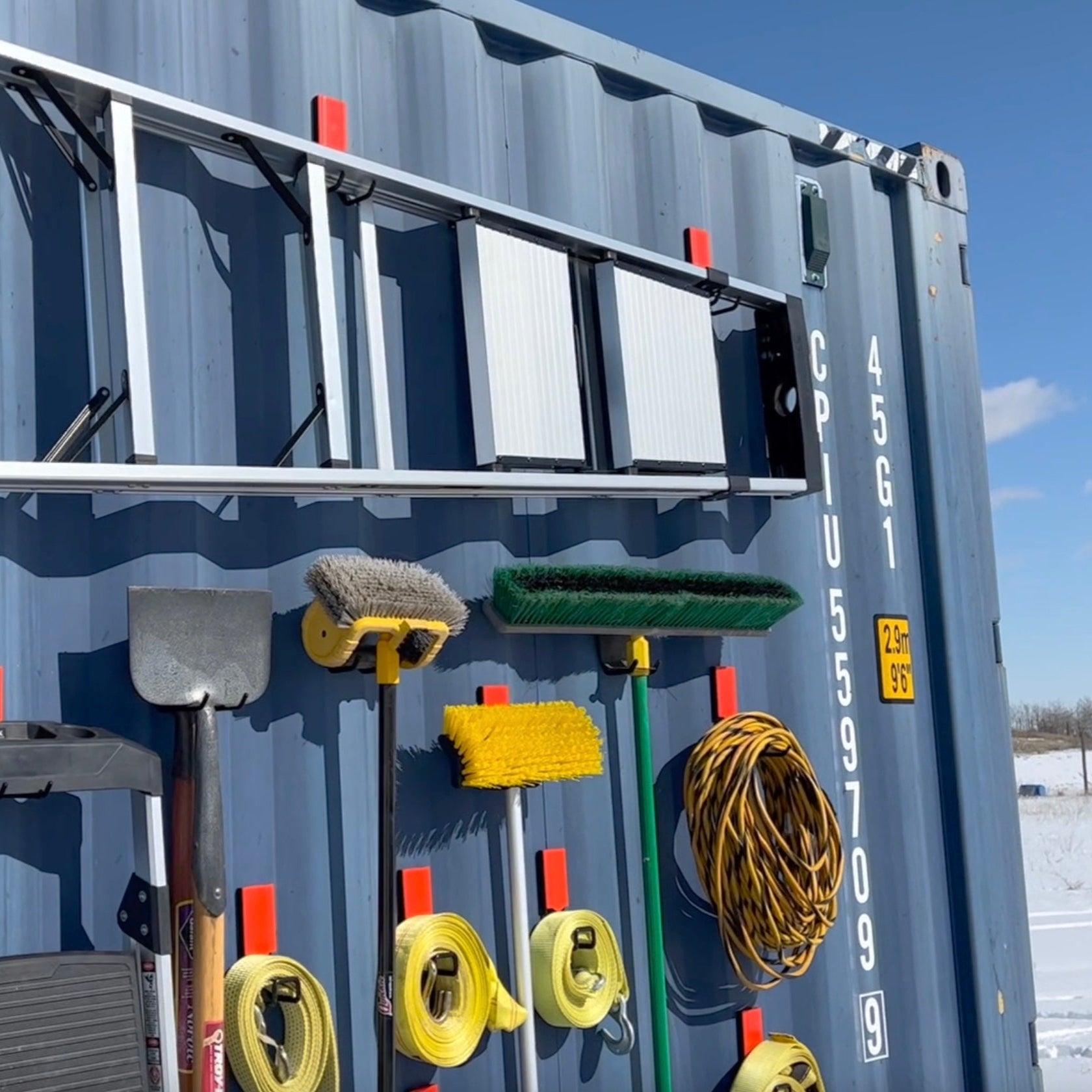 Gator Magnetics 4-inch Open Hook mounted on steel shipping container, holding ladder, push broom, snow brush, extension cord, and tie-down straps for outdoor storage.