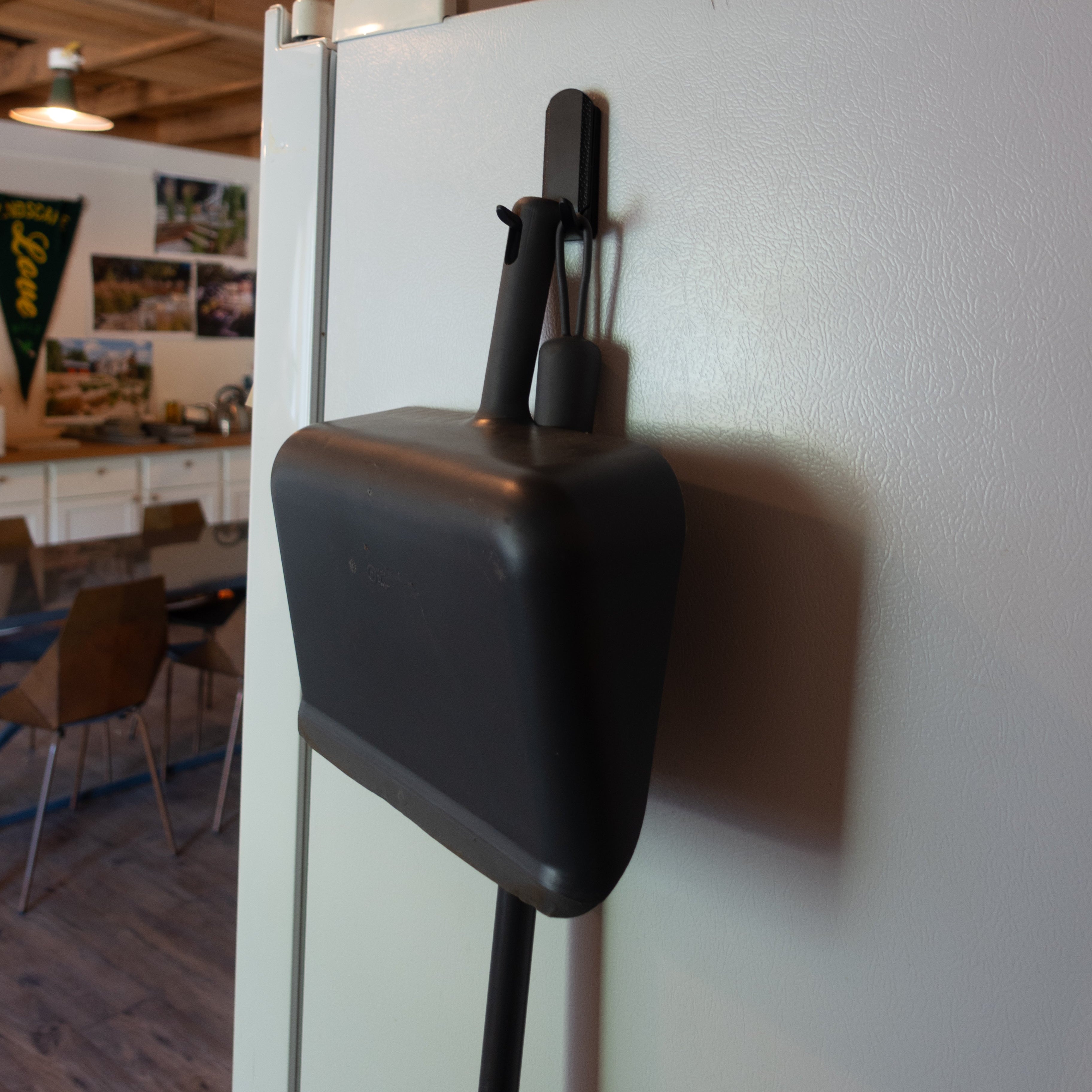 A black dustpan and brush are hung on a flat, white steel refrigerator door using a black Gator Magnetics magnetic hook, demonstrating easy, tool-free installation versus traditional screw in utility hooks.