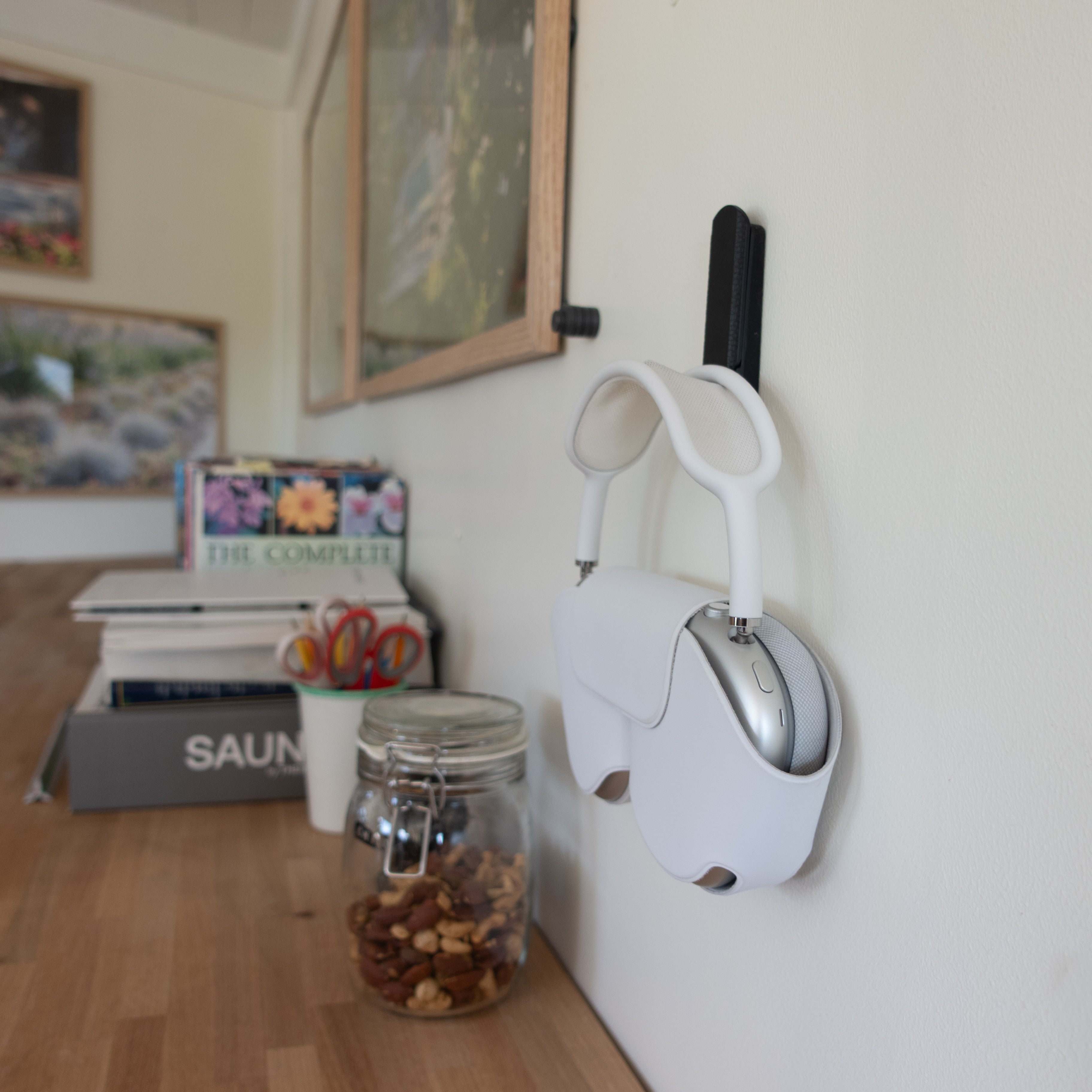 Black magnetic hook mounted on a white wall holding a pair of over-ear headphones in a white case, providing organized desk-side storage in a home office.