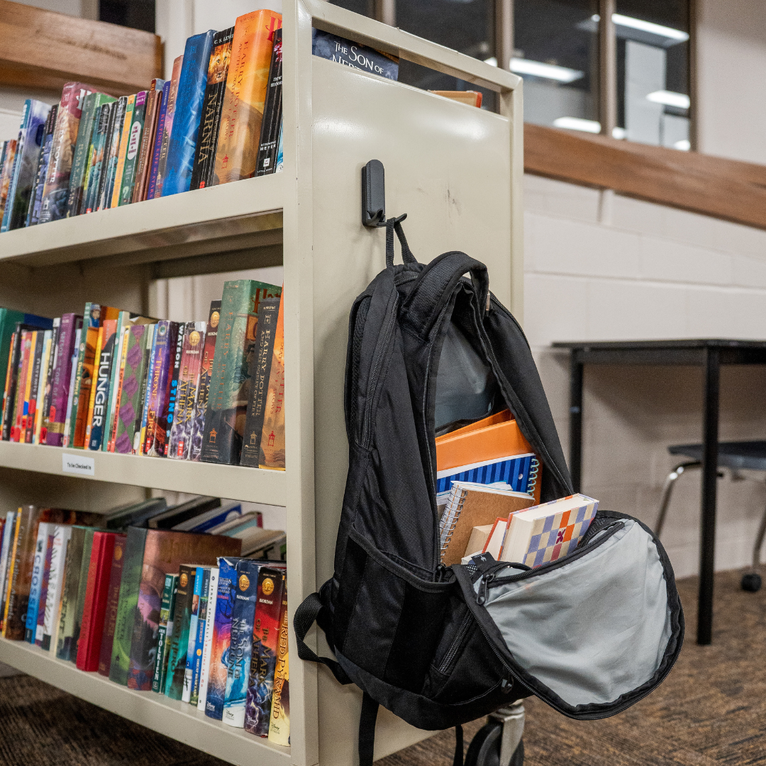 Black magnetic hook attached to a metal library bookshelf holding a black backpack filled with books and school supplies, providing practical storage in a public space.