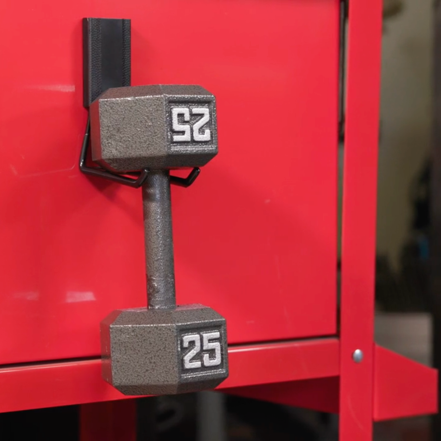 A 25-pound dumbbell hangs on a red tool cabinet steel surface using a Gator Magnetics red magnetic hook.