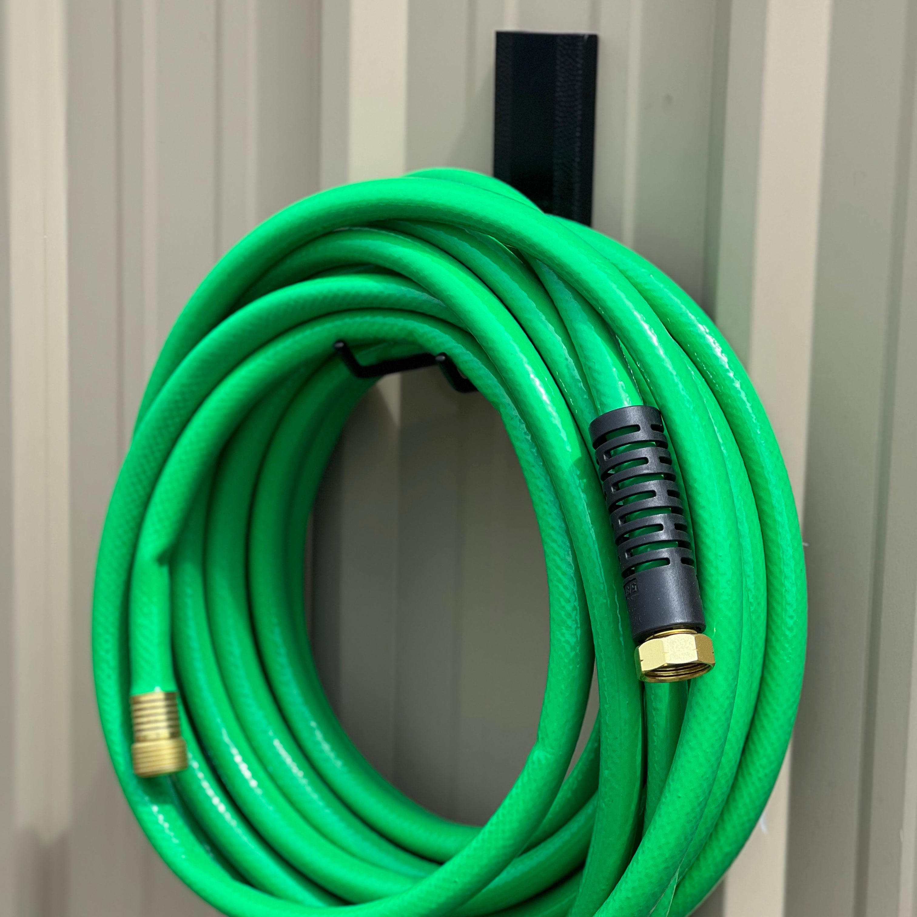 A green garden hose is neatly coiled and stored on a black magnetic hook attached to a vertical corrugated steel wall panel, demonstrating the strong magnetic capacity of Gator Magnetics products for organizing outdoor and workshop spaces.