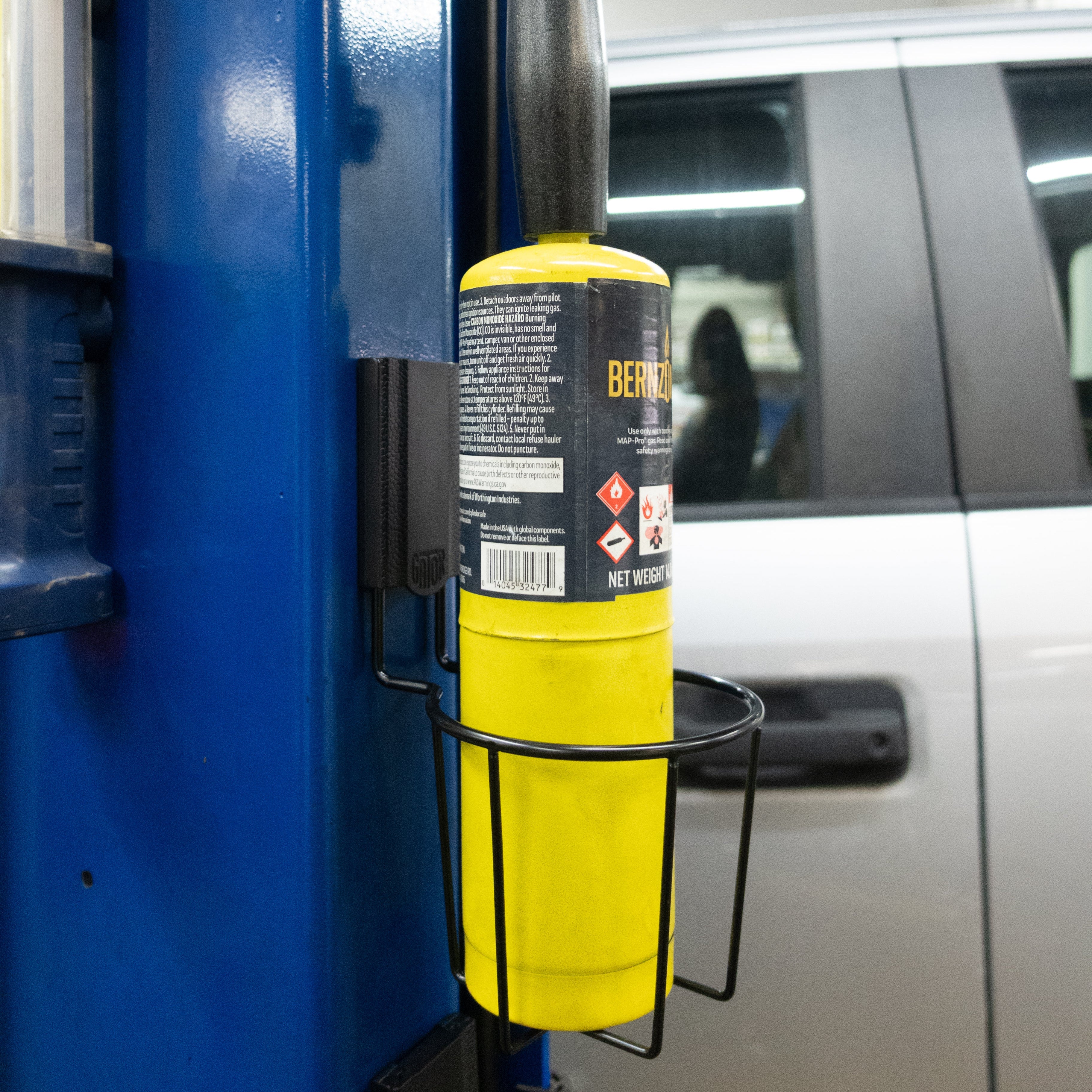 Auto shop storage and organization setup using a Gator Magnetics magnetic cup holder mounted on a blue automotive lift post, holding a yellow Bernzomatic torch cylinder, with a silver work van visible in the background.