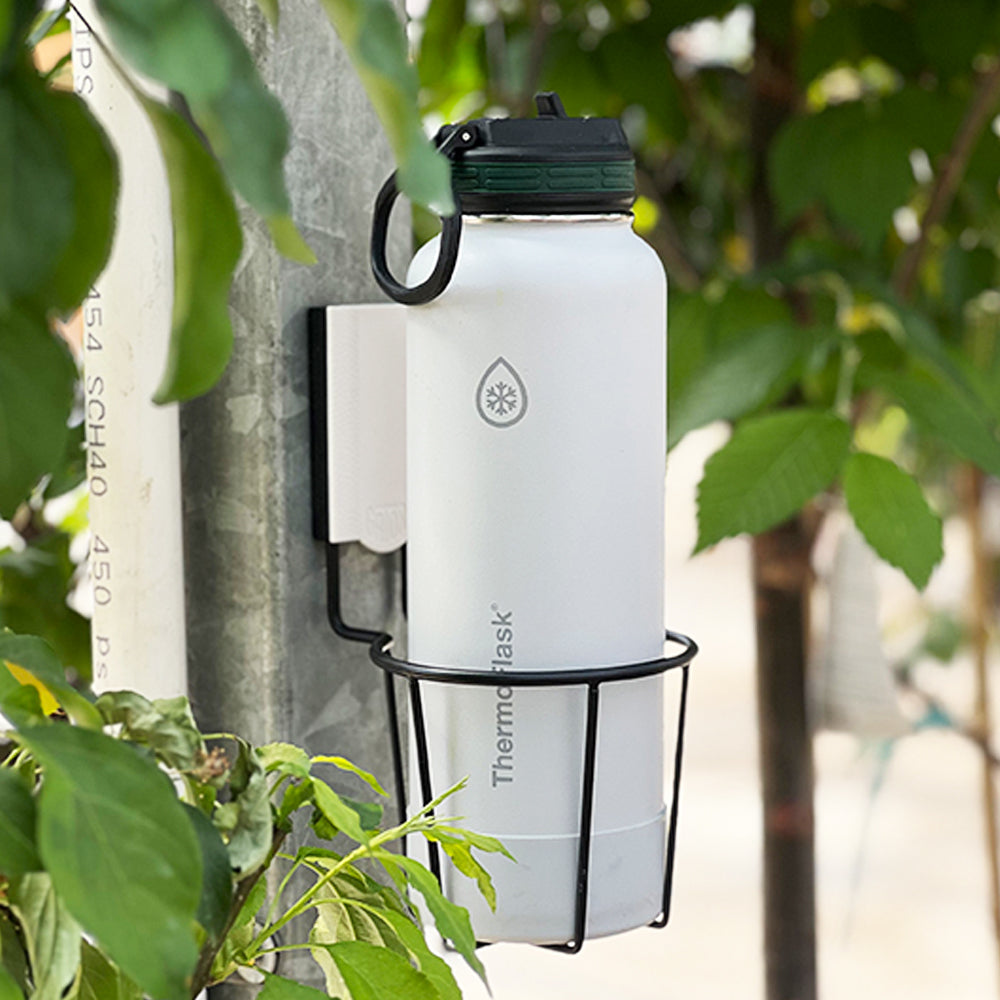 Greenhouse storage and organization setup with a Gator Magnetics magnetic cup holder mounted on a metal support post, holding a white ThermoFlask water bottle, surrounded by leafy plants and PVC irrigation pipes in the background.