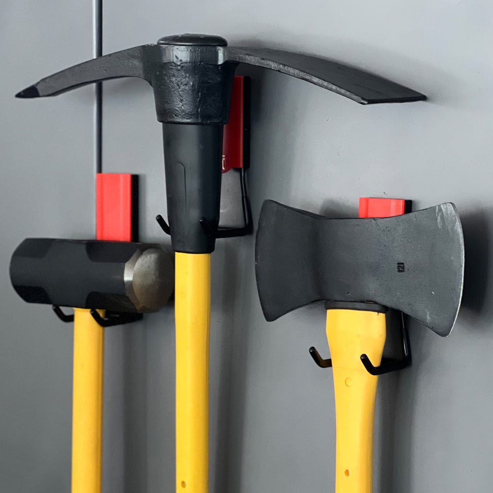 Three yellow-handled tools, a sledgehammer, pickaxe, and hatchet, are hung on red Gator Magnetics magnetic hooks attached to a flat, gray steel wall panel, demonstrating versatile heavy tool storage ideal for securing items like a leaf blower hook.