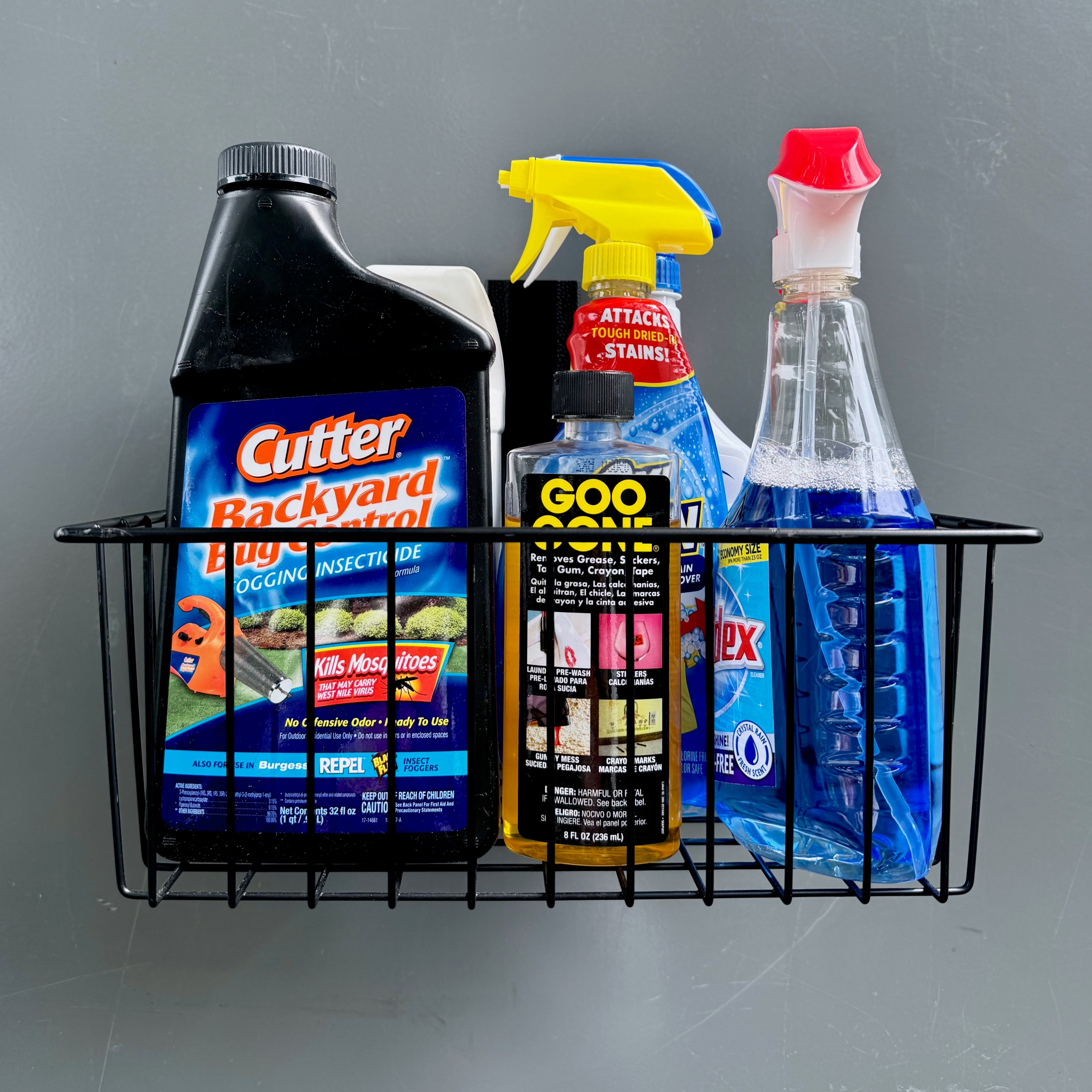 A black magnetic storage basket from Gator Magnetics holds insecticide, Goo Gone, Windex, and a blue cleaning spray hung on a flat gray steel wall panel, demonstrating heavy-duty, tool-free organization ideal for garages or sheds.
