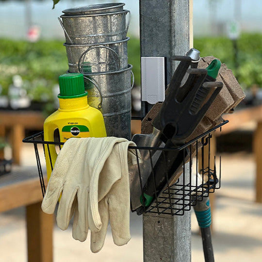 A black Gator Magnetics Magnetic Storage Basket is mounted to a flat galvanized steel pole barn steel wall, holding a pair of garden gloves draped over the edge, a yellow bottle of plant nutrients, three small galvanized metal buckets stacked inside, and garden tools with biodegradable seedling trays tucked inside, demonstrating effective use of heavy duty wall hooks 100 lbs for outdoor gardening organization.
