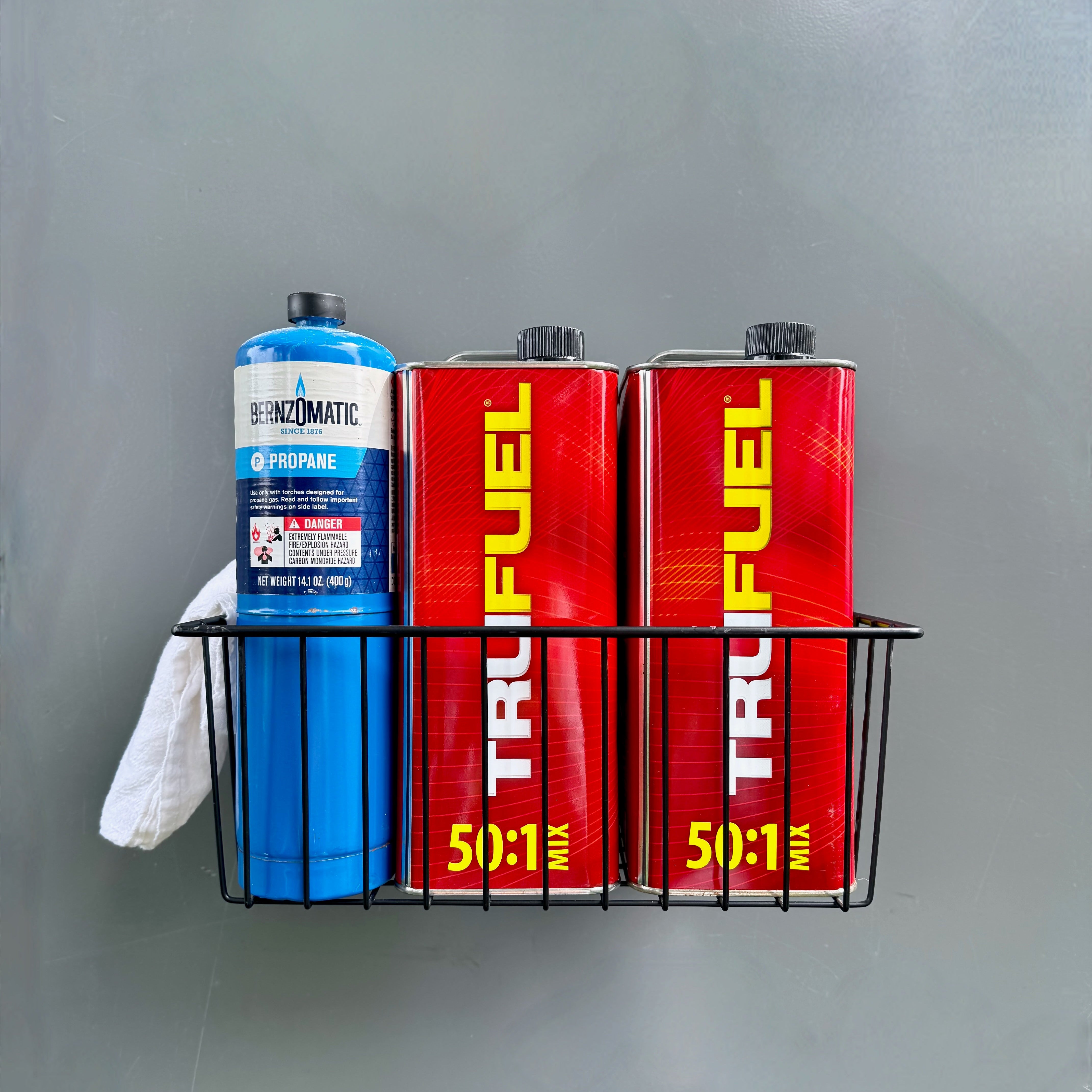 A black Gator Magnetics Magnetic Storage Basket holding one blue Bernzomatic propane cylinder, two red TruFuel fuel cans, and a white cloth, mounted on a flat steel wall panel.