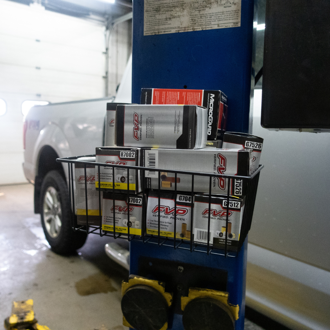 Gator Magnetics magnetic basket mounted on a blue steel beam in an automotive shop, providing storage and organization for multiple boxed oil filters, keeping parts easily accessible next to a vehicle lift with a pickup truck in the background.