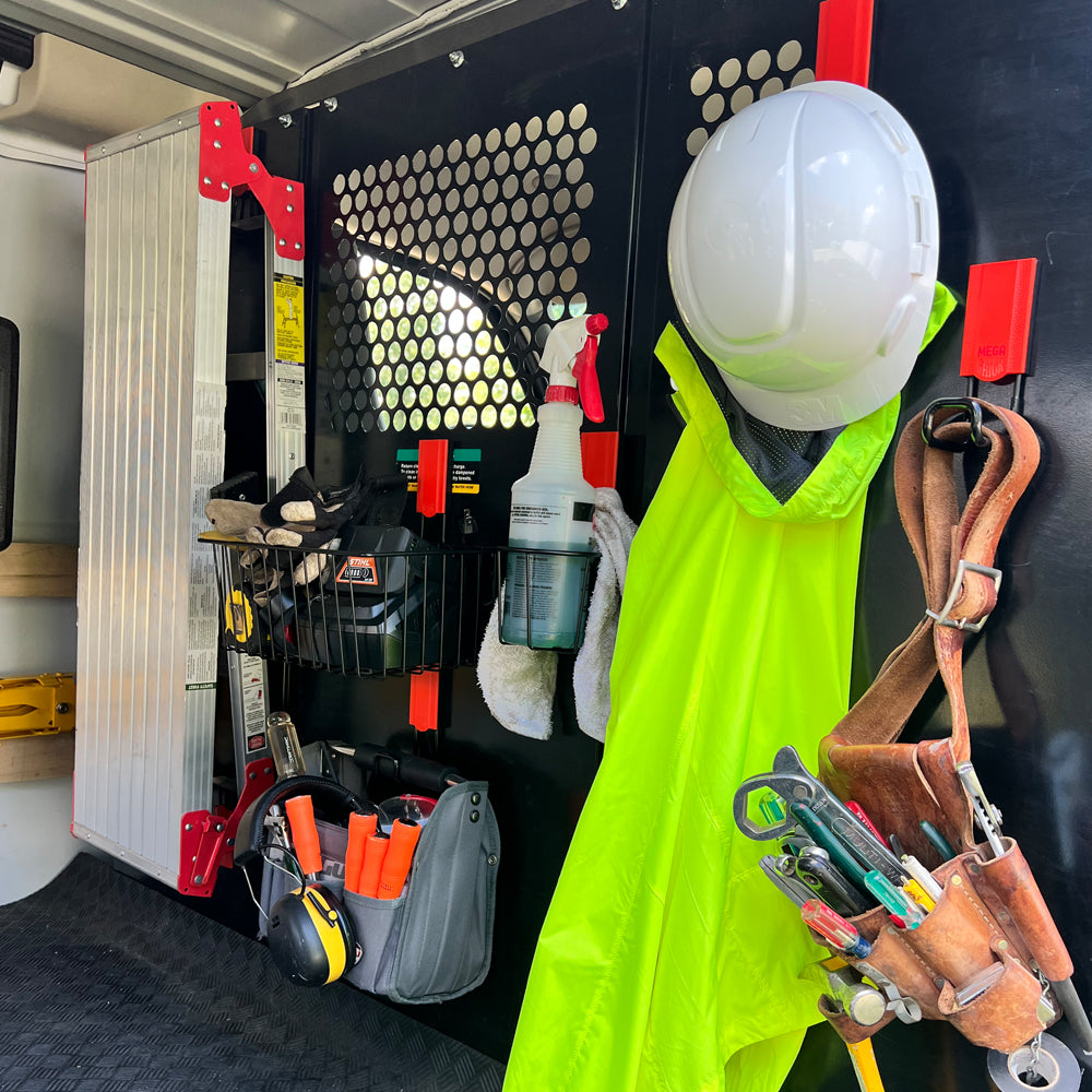 A black steel wall panel inside a work van organized with red Gator Magnetics magnetic hooks and a magnetic storage basket holding gloves, a battery charger, and a spray bottle, while a high-visibility vest and white hard hat hang nearby and a leather tool belt with hand tools is also stored, showcasing efficient garage organization wall systems.