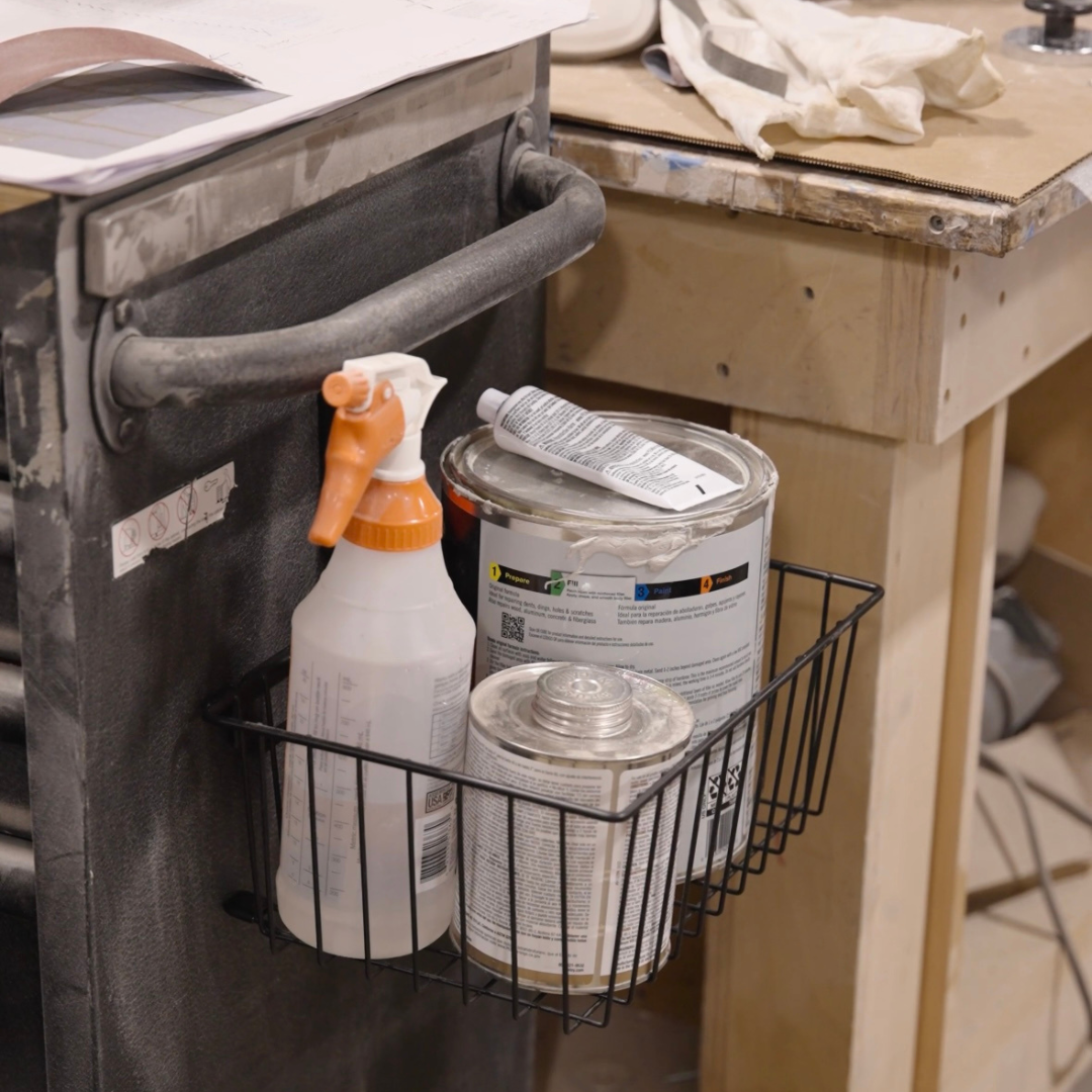 Gator Magnetics heavy-duty magnetic basket attached to a metal tool cart in a workshop, providing storage and organization for a spray bottle, paint cans, and woodworking supplies, keeping tools and materials off the workbench and within easy reach.