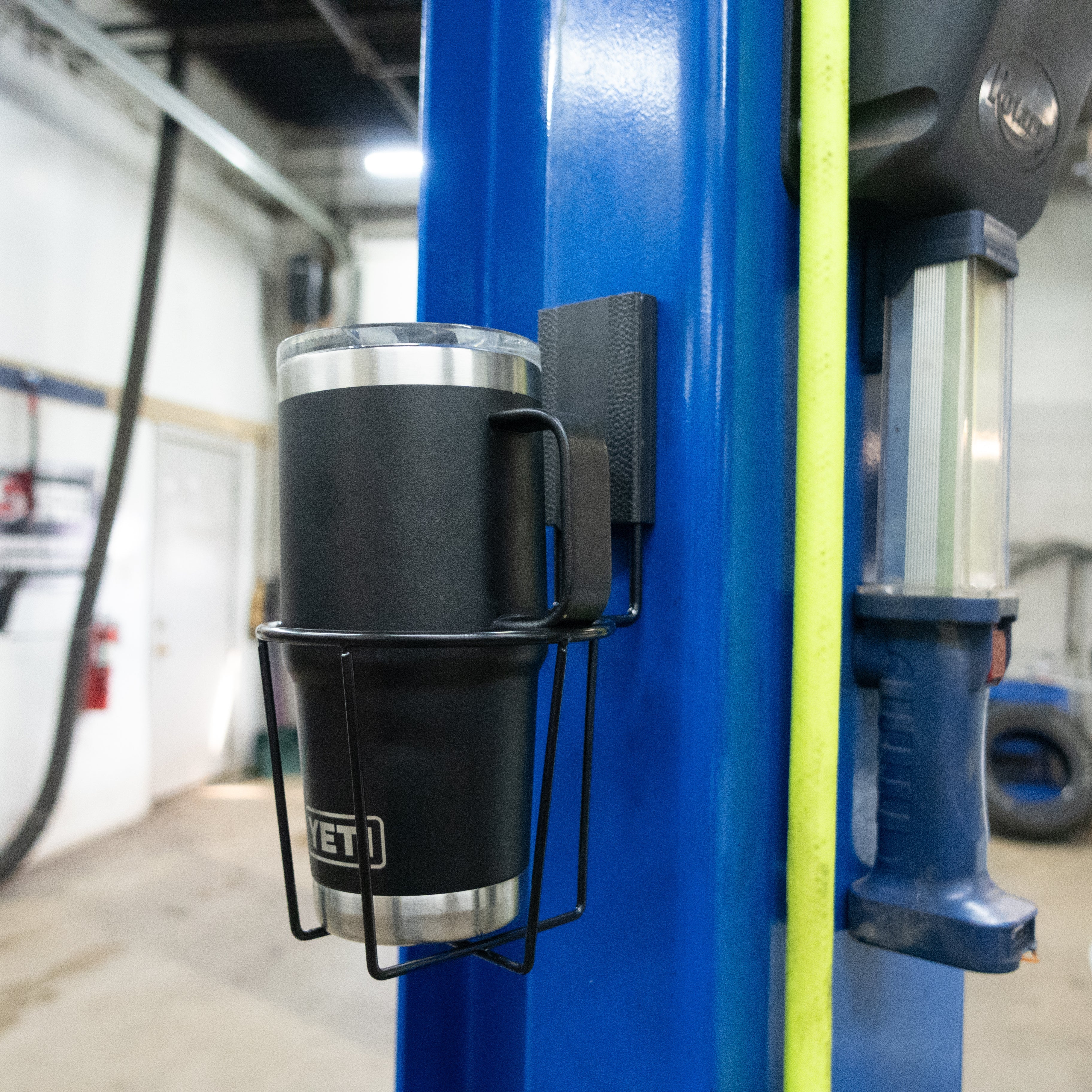 Gator Magnetics magnetic cup holder for auto shop storage and organization, mounted on a blue automotive lift holding a black YETI tumbler, with tools, equipment, and a garage workspace visible in the background.