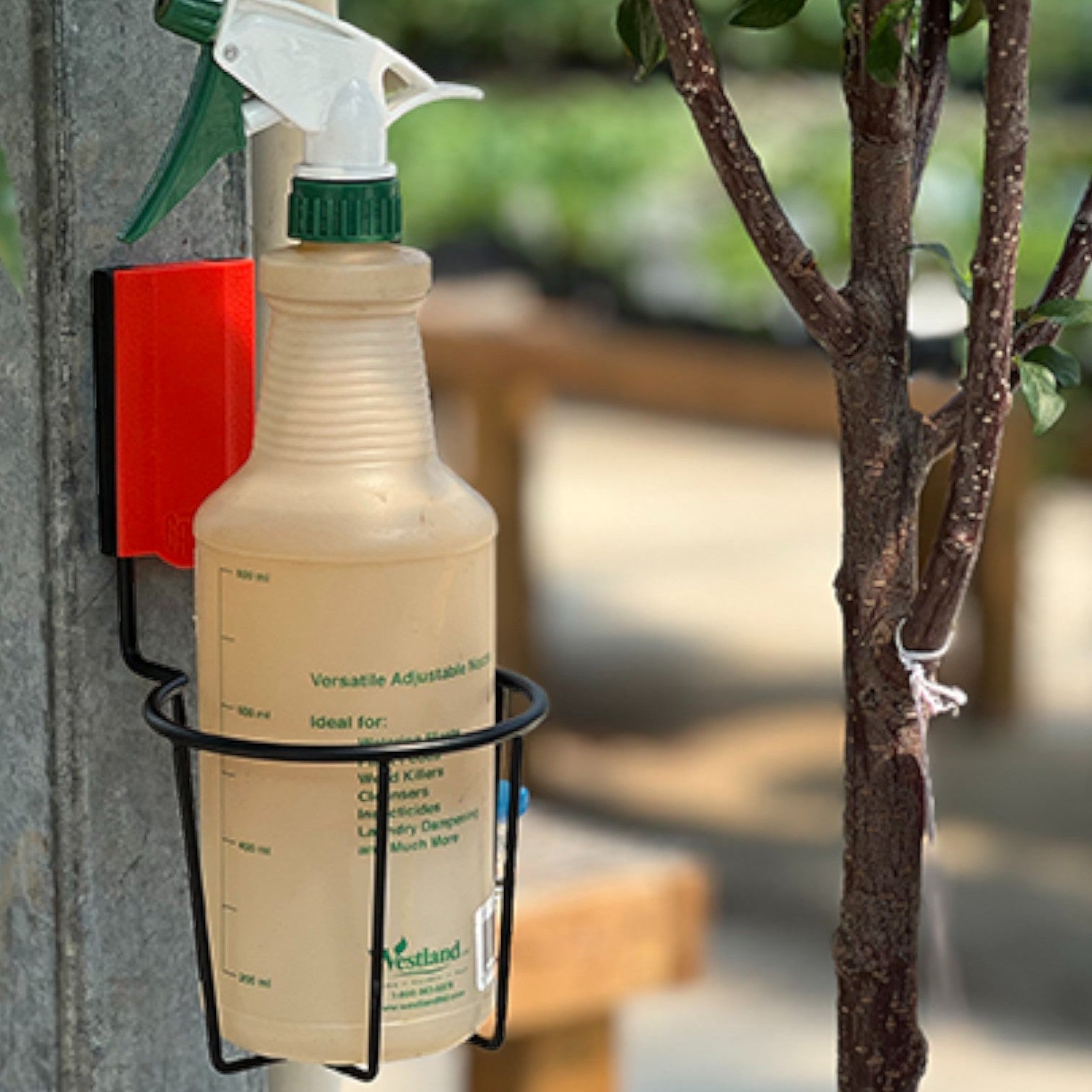 Gator Magnetics magnetic cup holder for garden storage and organization, attached to a metal post holding a large spray bottle, with plants, a small tree, and outdoor gardening area in the background.