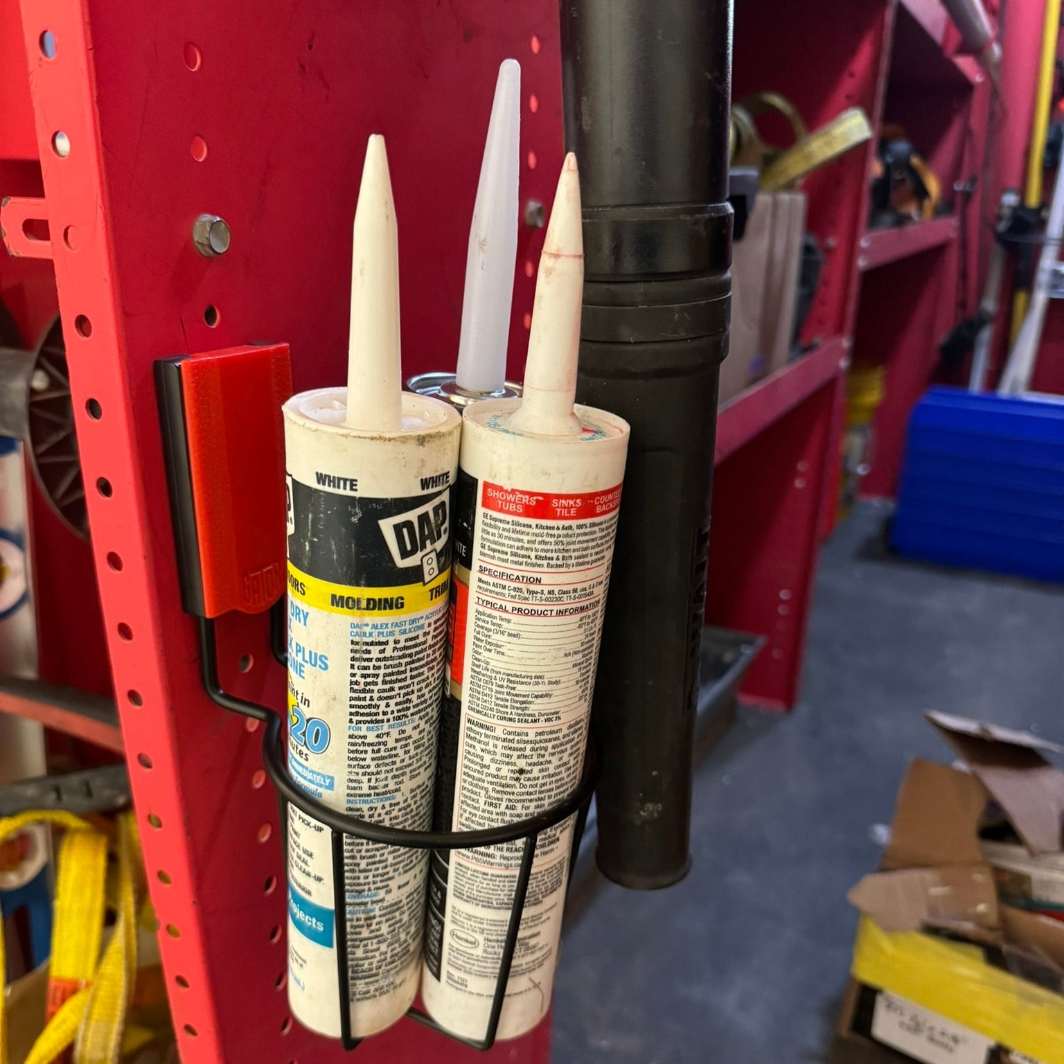 Gator Magnetics magnetic cup holder for utility van storage and organization, mounted on a red metal shelving unit holding two caulk tubes and a flashlight, with organized tools, storage bins, and shelves visible in the background.