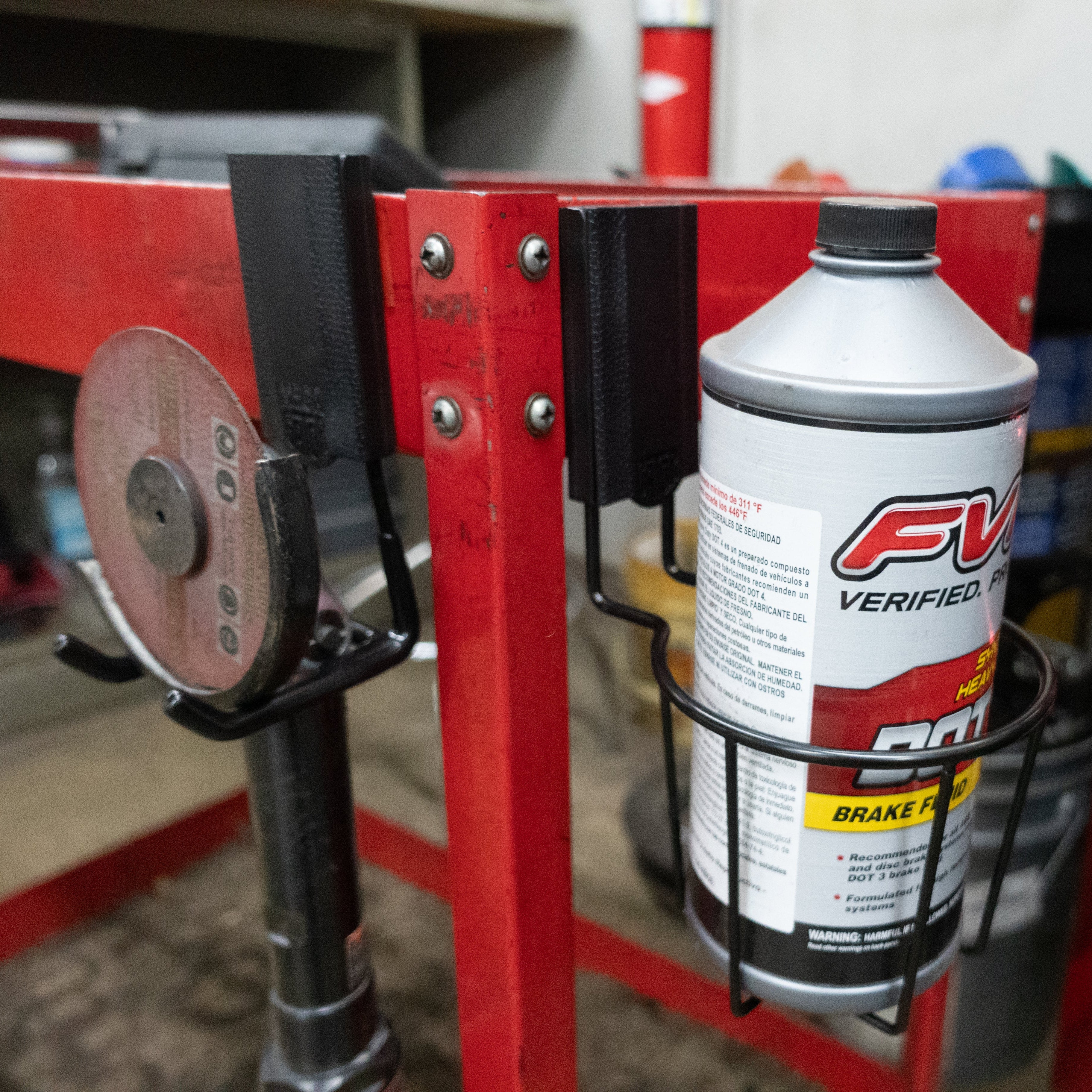 Gator Magnetics magnetic cup holder for workshop storage and organization, attached to a red metal workbench to hold a brake fluid can securely, alongside a magnetic hook holding a grinding wheel, keeping tools and supplies neatly arranged.