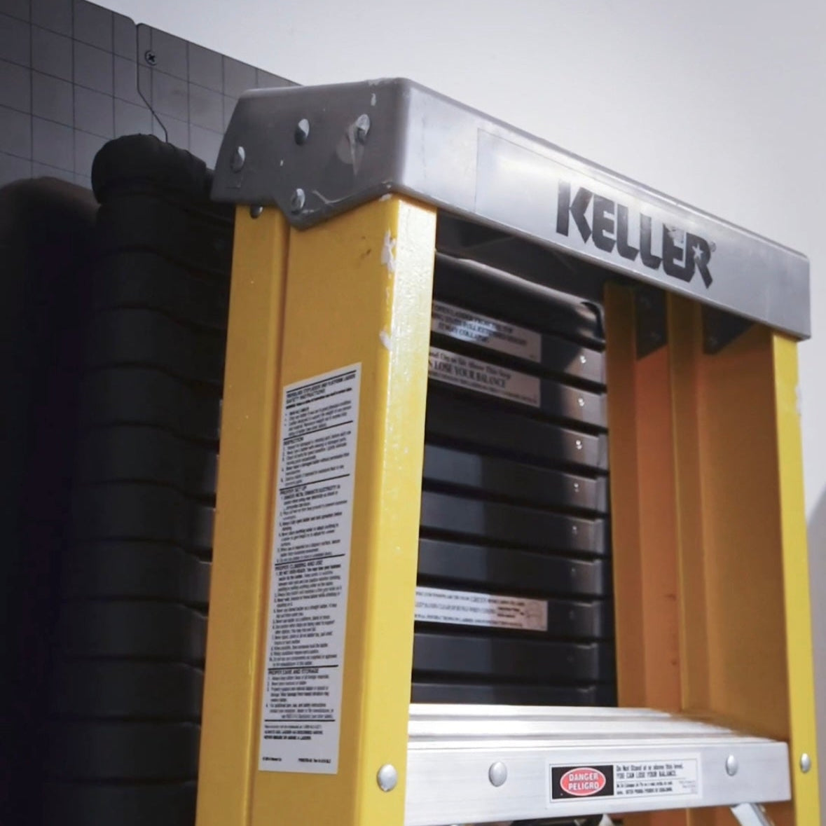 Gator Magnetics heavy duty magnetic hook mounted on a black steel wall storage system in a garage holding a yellow Keller step ladder for secure garage storage and organization, with additional wall-mounted equipment visible in the background.