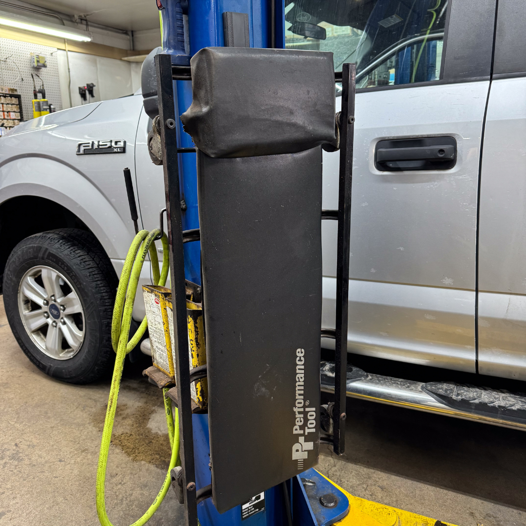 A padded black tool holder with "Performance Tool" branding is mounted on a blue steel pole in a garage, with a coiled yellow air hose hanging from the frame, positioned next to a white Ford F-150 truck.