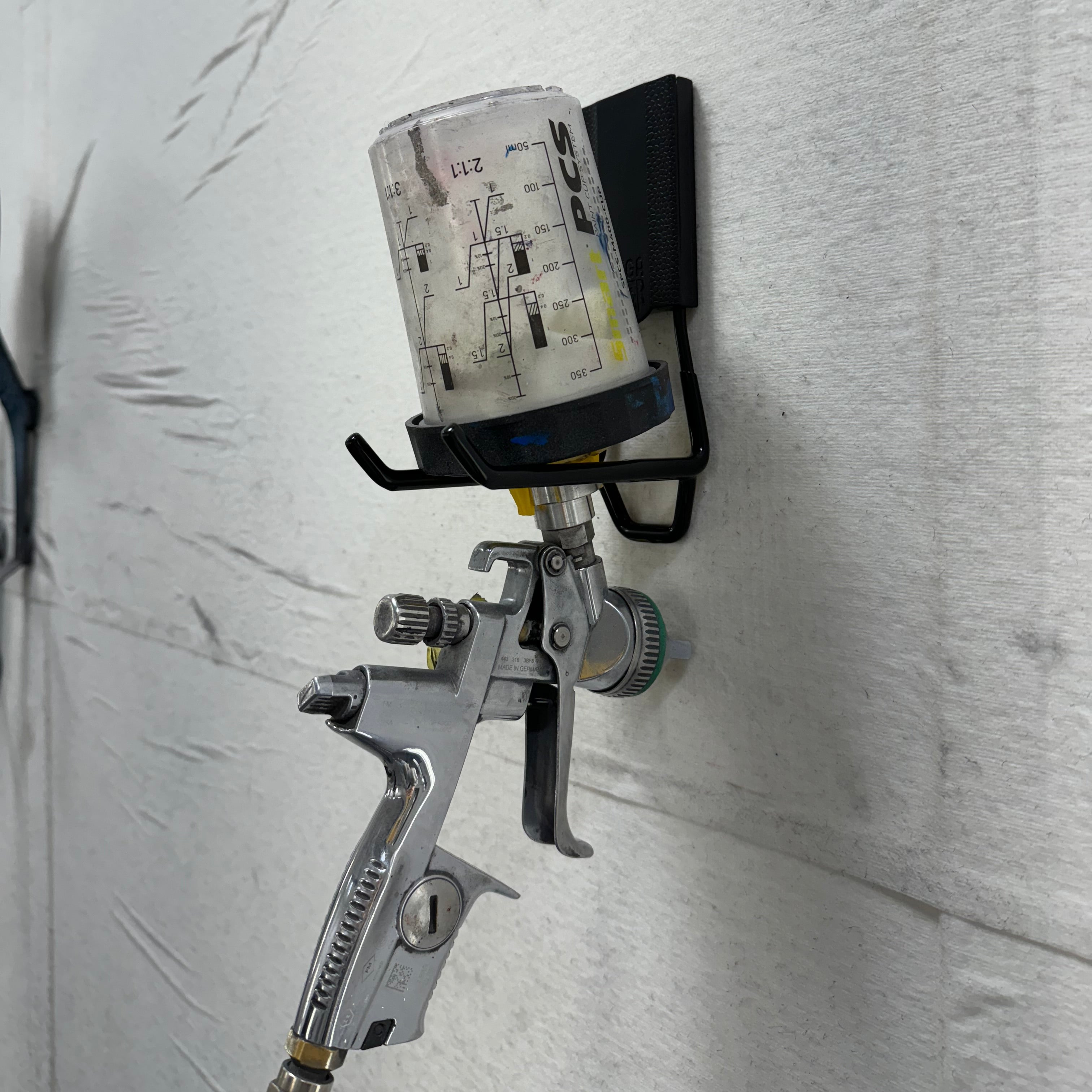 Gator Magnetics magnetic hook securing automotive paint spray gun with mixing cup on steel wall in body shop for organized storage.