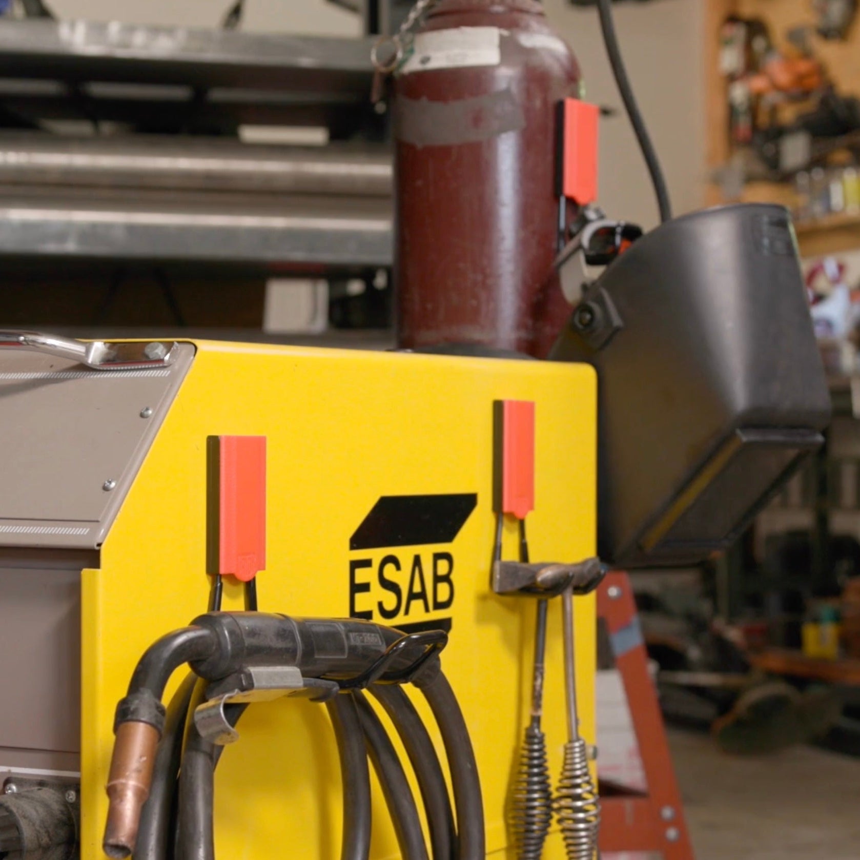 Gator Magnetics heavy duty magnetic hooks holding welding torch cables on an ESAB welder in a workshop, with welding helmet and gas cylinder in the background for organized tool storage and improved workspace organization.