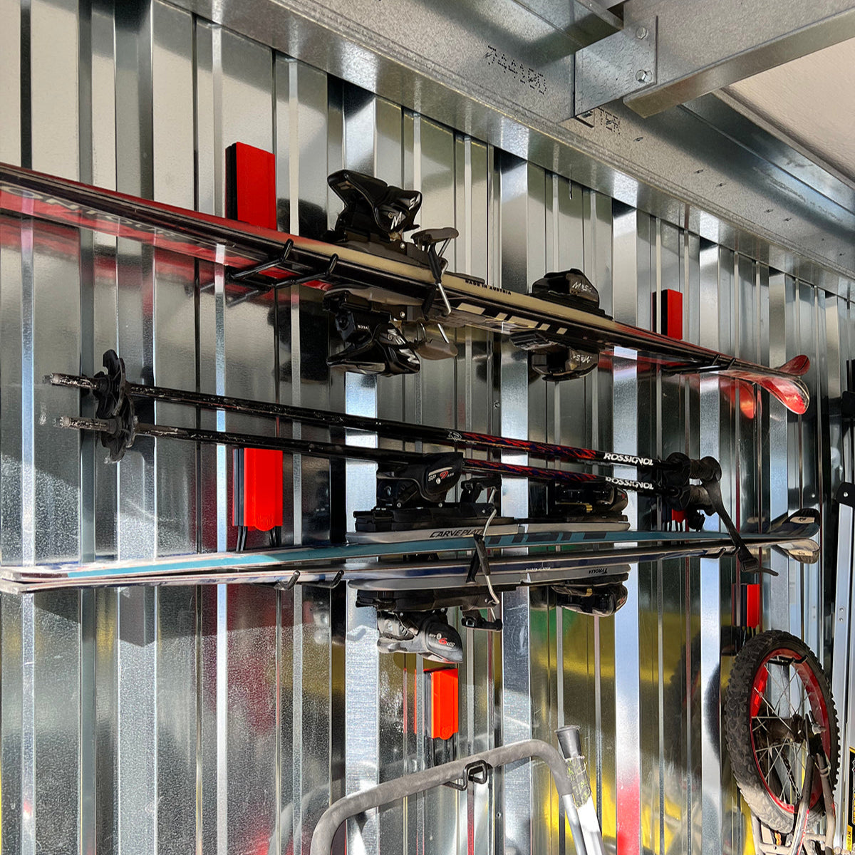 Two pairs of skis and two pairs of ski poles are neatly held on bright red Gator Magnetic Hooks mounted on a shiny, corrugated steel pole barn wall, demonstrating efficient winter gear and backpack storage home organization.