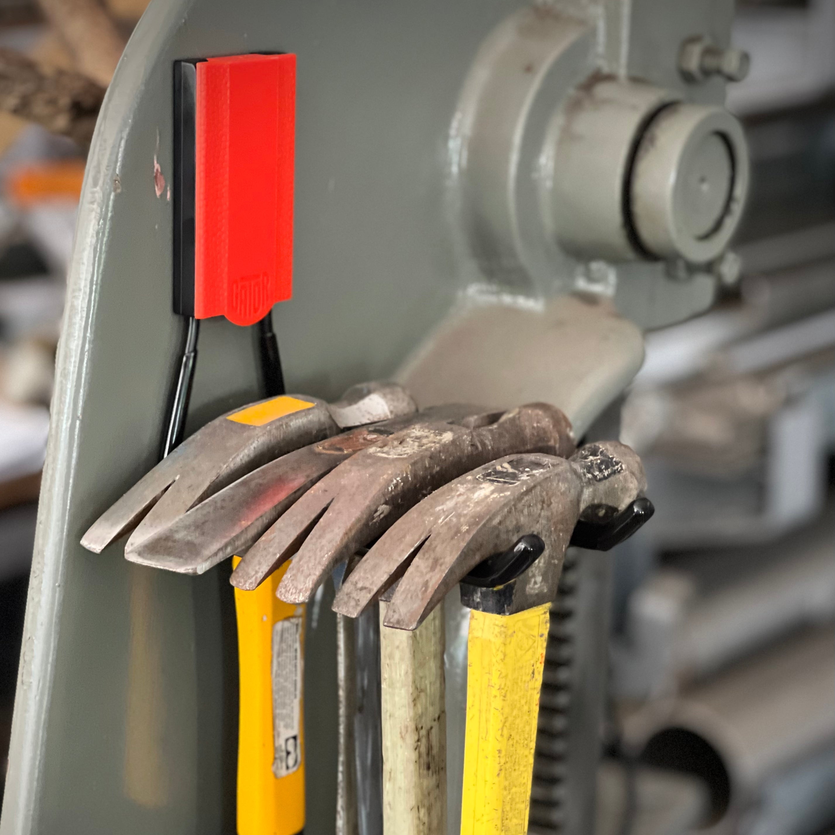 A red Gator Magnetics magnetic hook holds four claw hammers by their heads on a steel industrial machine surface.
