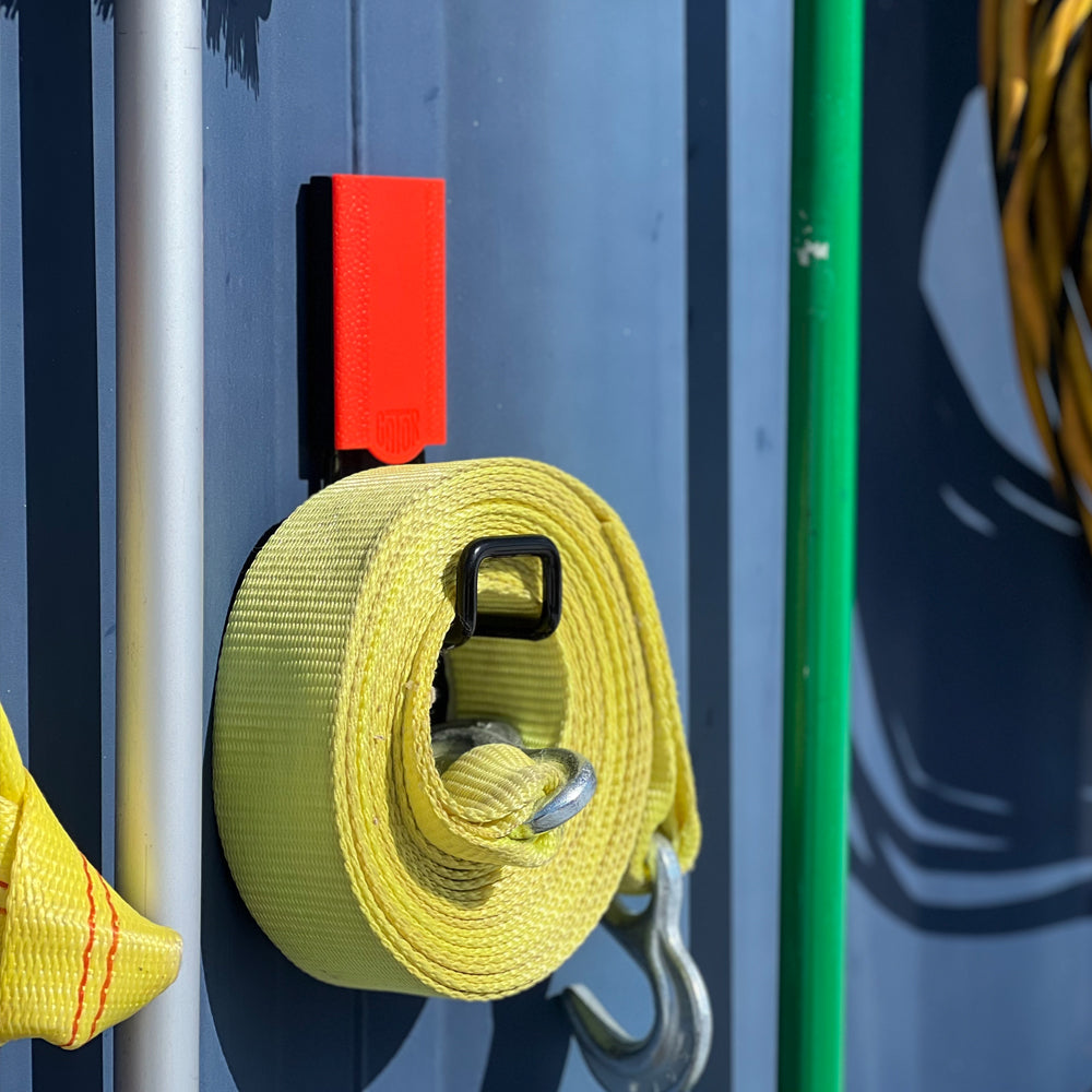 Rolled yellow ratchet strap hanging from a red Gator magnetic hook on a steel shipping container.