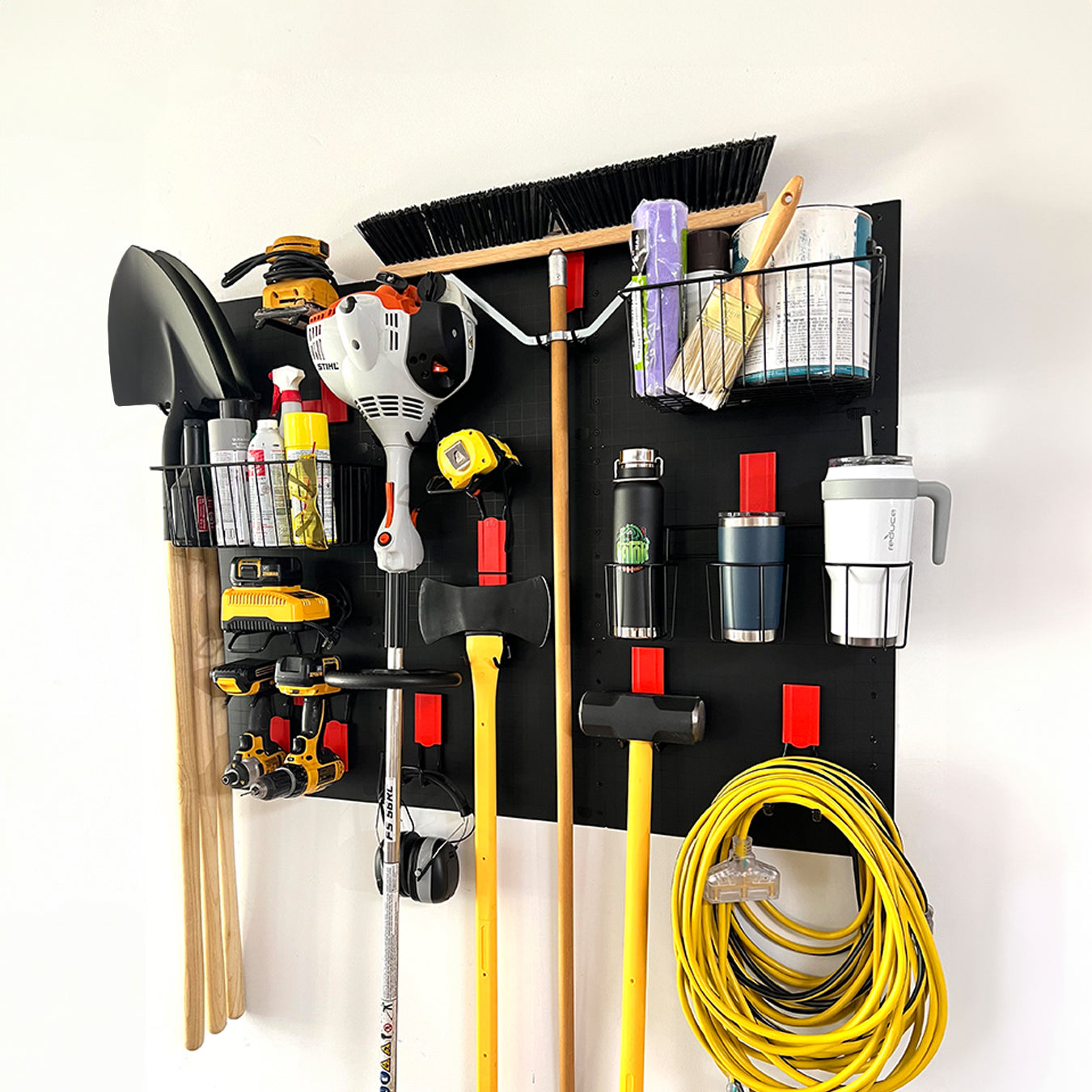 A black Gator Steel Wall Panel mounted on a white wall holds an assortment of tools and supplies including a yellow power drill, a white and orange string trimmer, a yellow sledgehammer and axe, a long-handled broom, two wire baskets with paint cans and brushes, several spray cans, a rolled purple tarp, three insulated drink tumblers, and coiled yellow extension cords, showcasing efficient storage for golf clubs and workshop equipment with red Gator Magnetic Hooks and baskets.