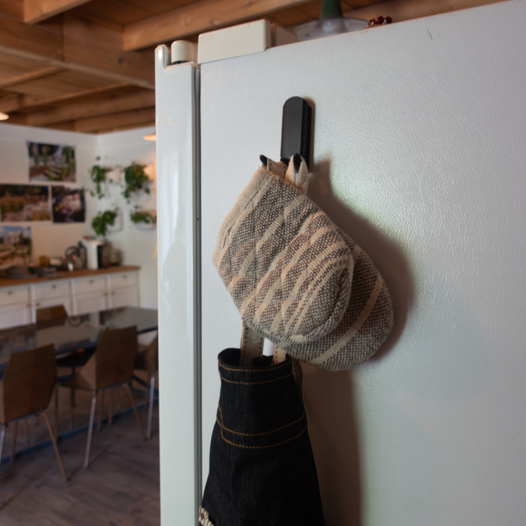 A striped oven mitt and a black apron are hung on a black magnetic hook attached to a white refrigerator door.