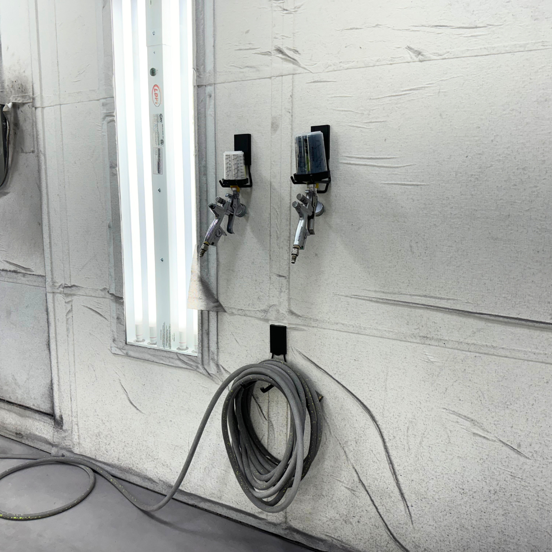 Two paint spray guns and a coiled air hose hang from black magnetic hooks on a steel garage wall panel.