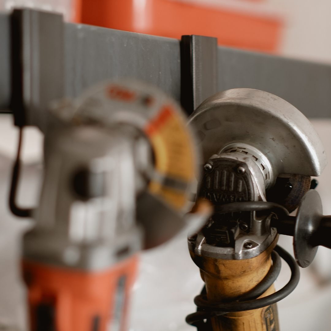 Two power angle grinders are hung on a horizontal steel rail using red magnetic hooks in a workshop setting.
