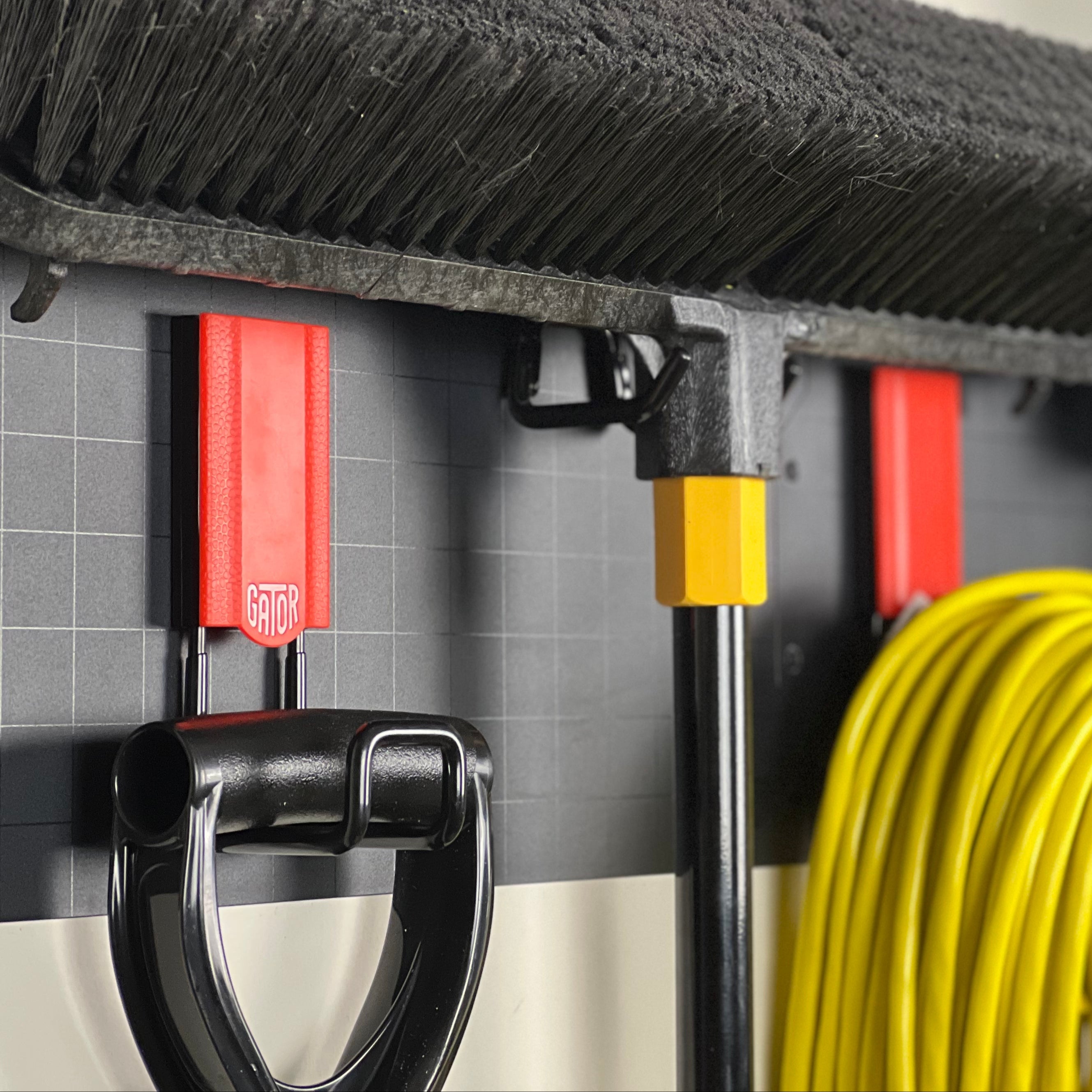 Two red Gator magnetic hooks hold a broom and yellow-handled tool on a black steel wall panel.