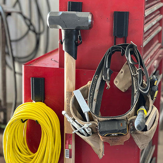 Two red Gator Magnetics magnetic hooks hold a sledgehammer and a tool belt with various tools on the steel side panel of a red tool chest.