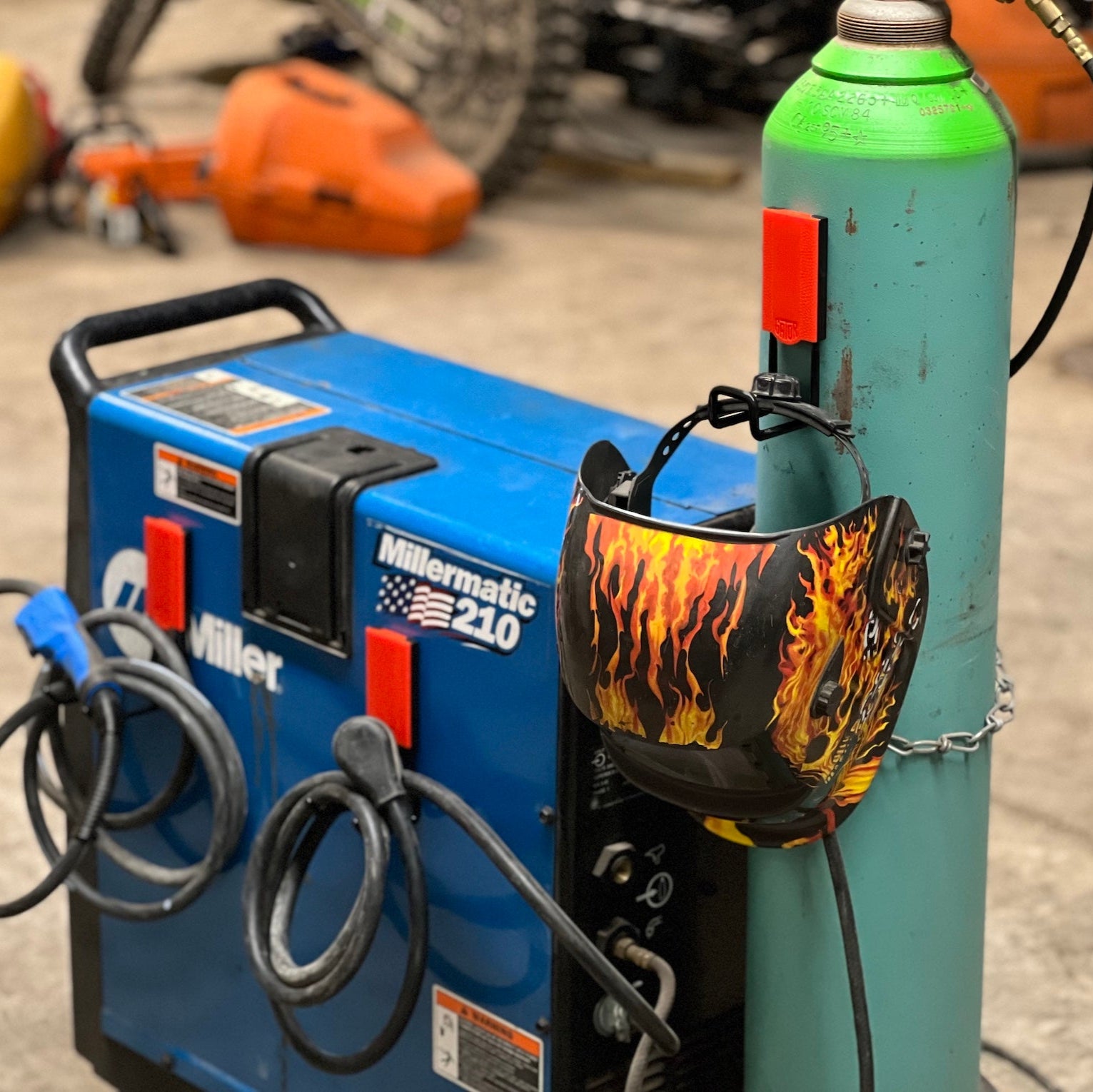 A welding helmet with flame decals hangs from a red Gator Magnetics magnetic hook attached to a green steel cylinder.