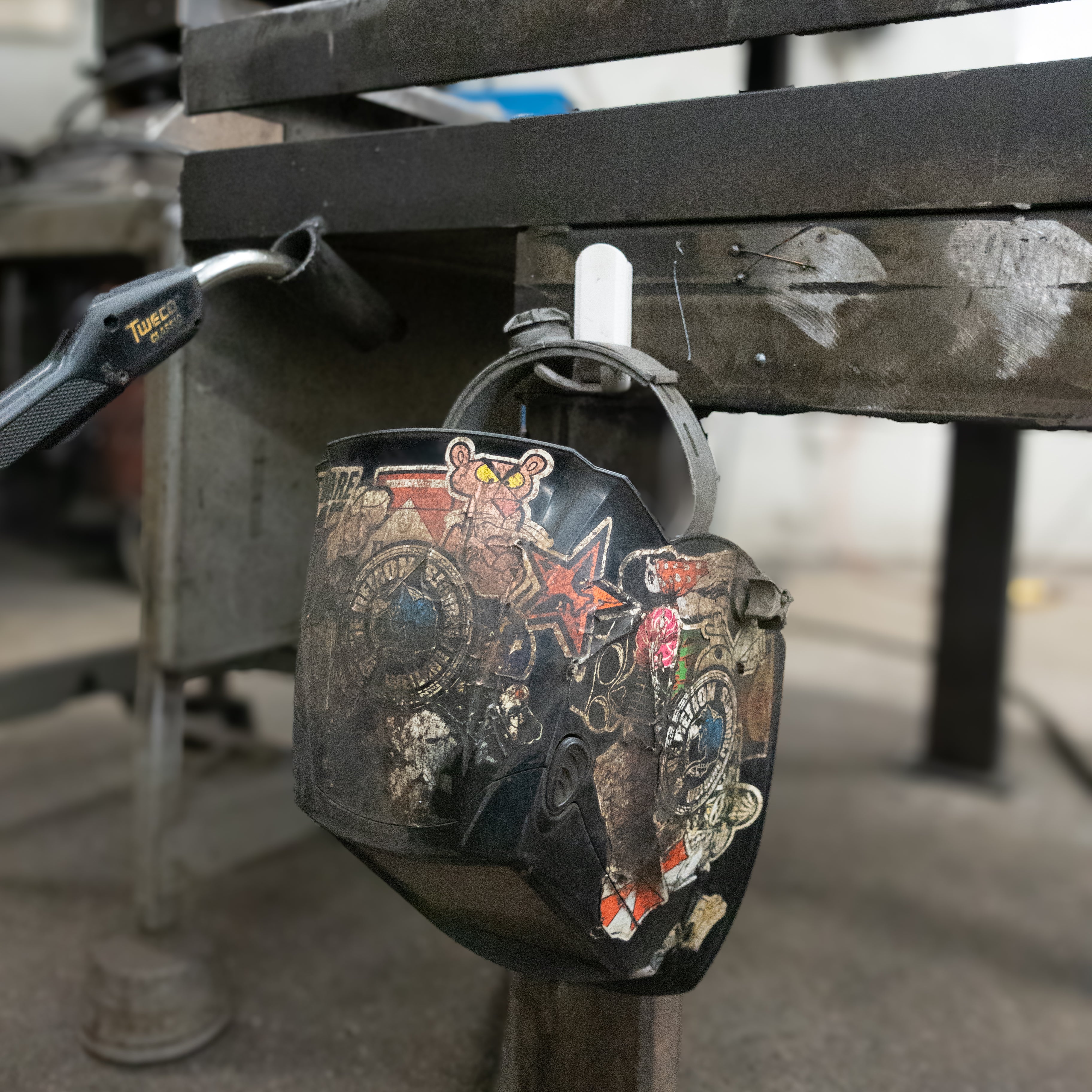 A welding helmet with various stickers is hanging from a red hook attached to a heavy-duty steel workshop table frame.