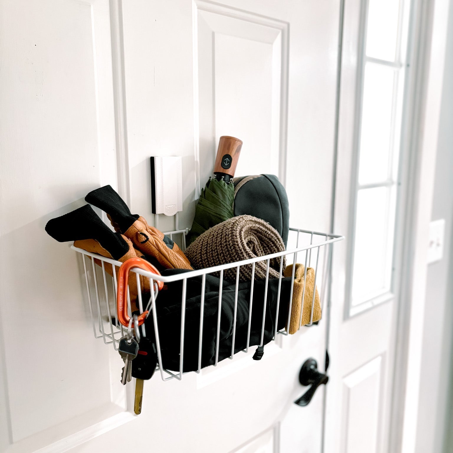 White magnetic storage basket on a steel door holding gloves, a scarf, umbrella and hat.