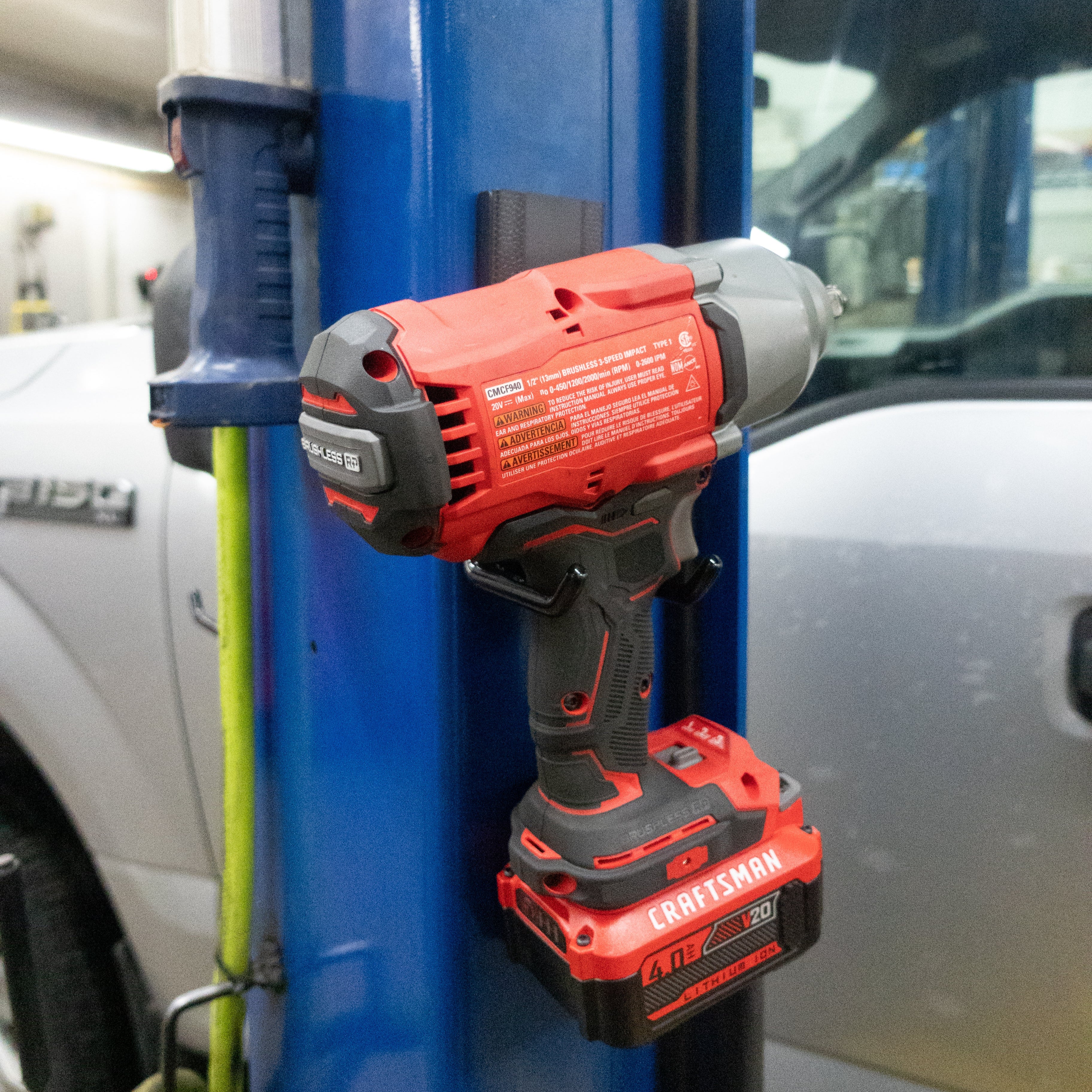 A red and black cordless Craftsman impact driver is hanging by its handle on a red Gator Magnetics magnetic hook attached to a blue steel lift post in a workshop environment, showcasing effortless tool storage on a flat steel surface.