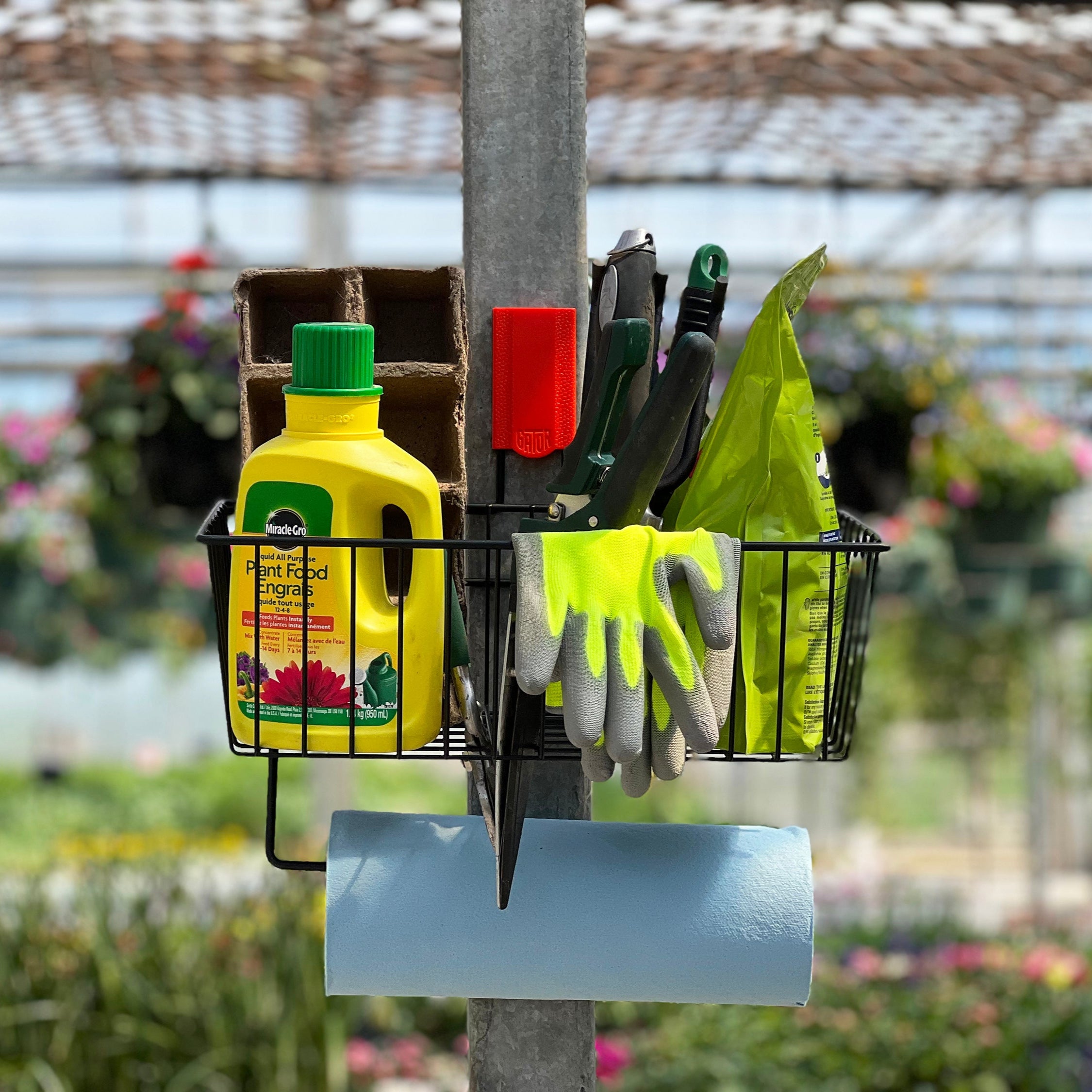 A black magnetic storage basket from Gator Magnetics holding a yellow bottle of plant food, gardening gloves, a green zip tie, pruning shears, and a green packet, mounted on a vertical galvanized steel pole with a blue paper towel roll hanging below, demonstrating practical organization for gardening tools and suggesting where to store backpacks in similar compact spaces.