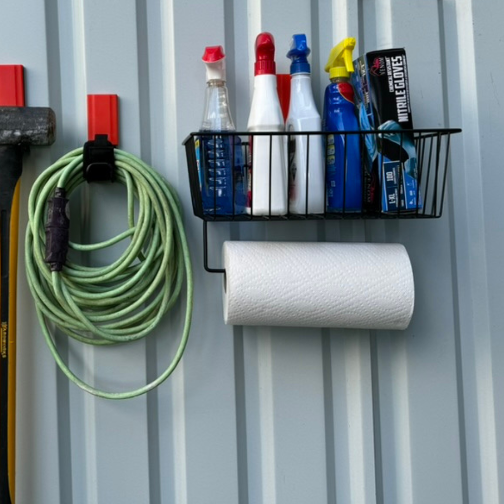 Magnetic basket for pole barn storage and organization, holding cleaning supplies, spray bottles, and nitrile gloves with a paper towel roll below, alongside magnetic hooks storing an extension cord and sledgehammer on a corrugated steel wall.